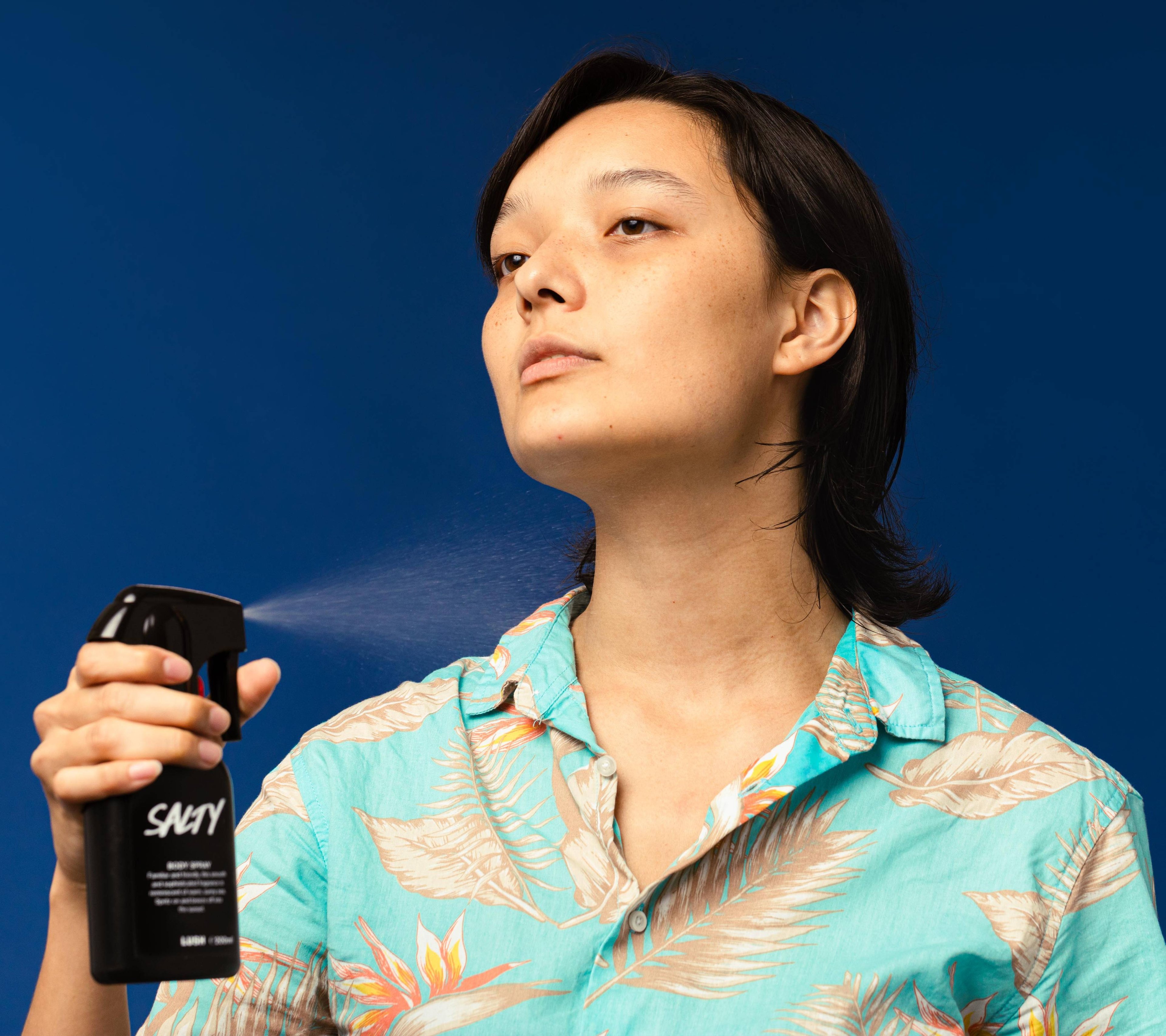 A person wearing a summery shirt sprays Salty body spray onto their neck and chest, looking blissed out and carefree.