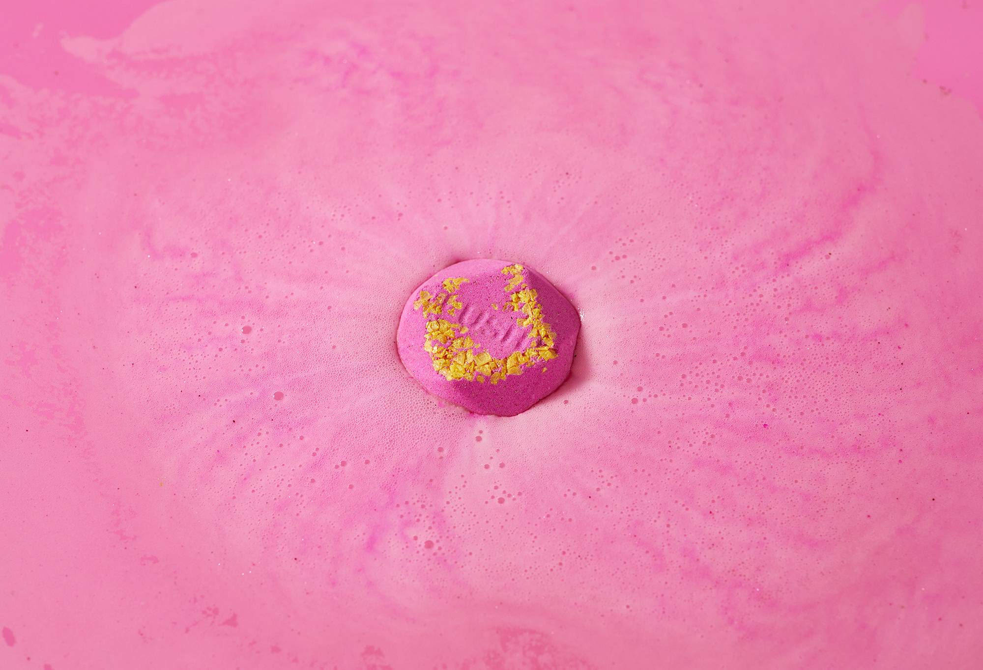 San San bath bomb sits on a sea of hot pink swirling foam with small pressed yellow-coloured salt and "Lush" embossed on top.