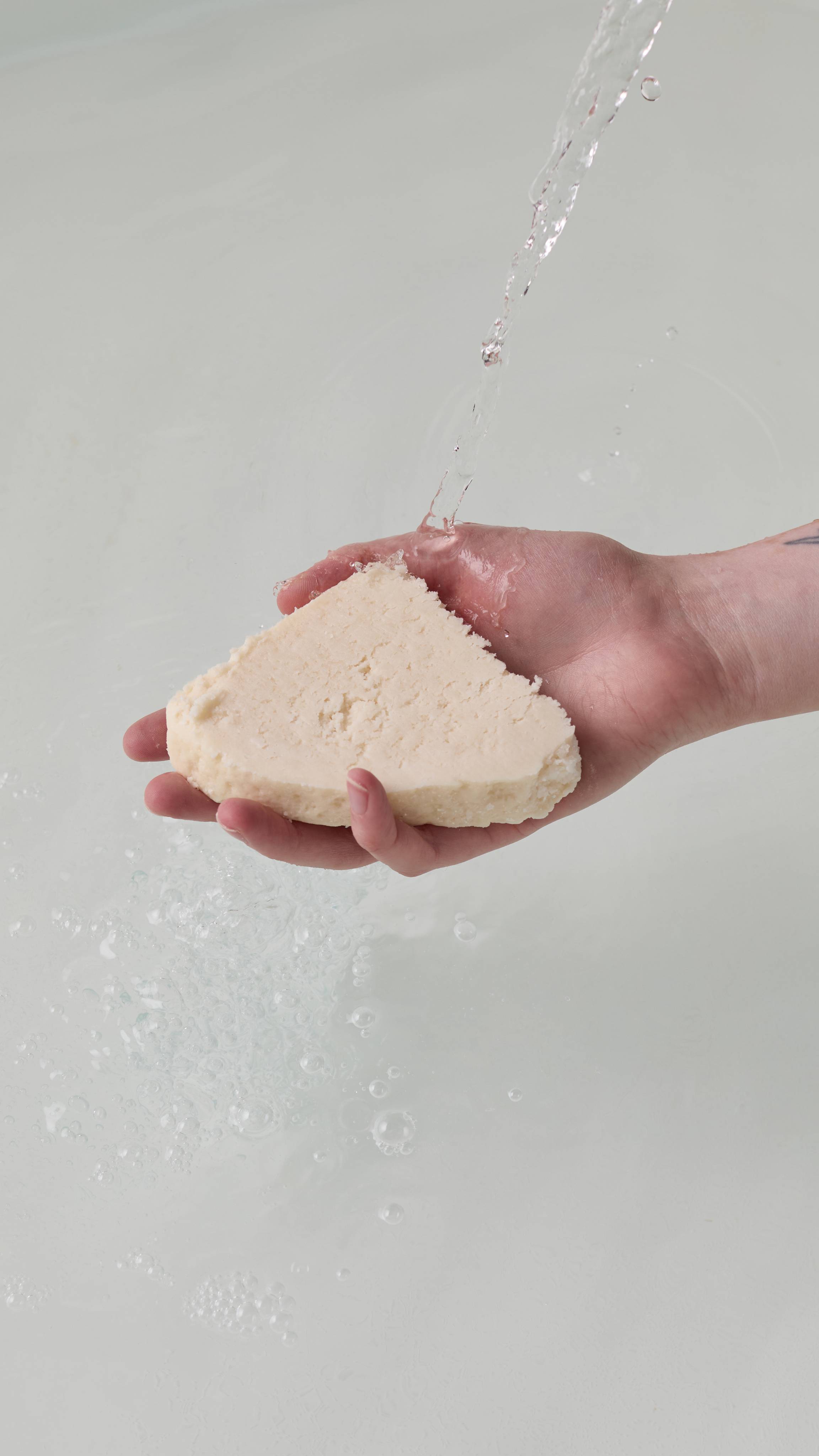 The image shows the model's hand reaching in from the right as it holds the creamy, crumbly Santa Moritz bubble bar under running tap water in a white porcelain bath. The water has a delicate layer of bubbles forming.