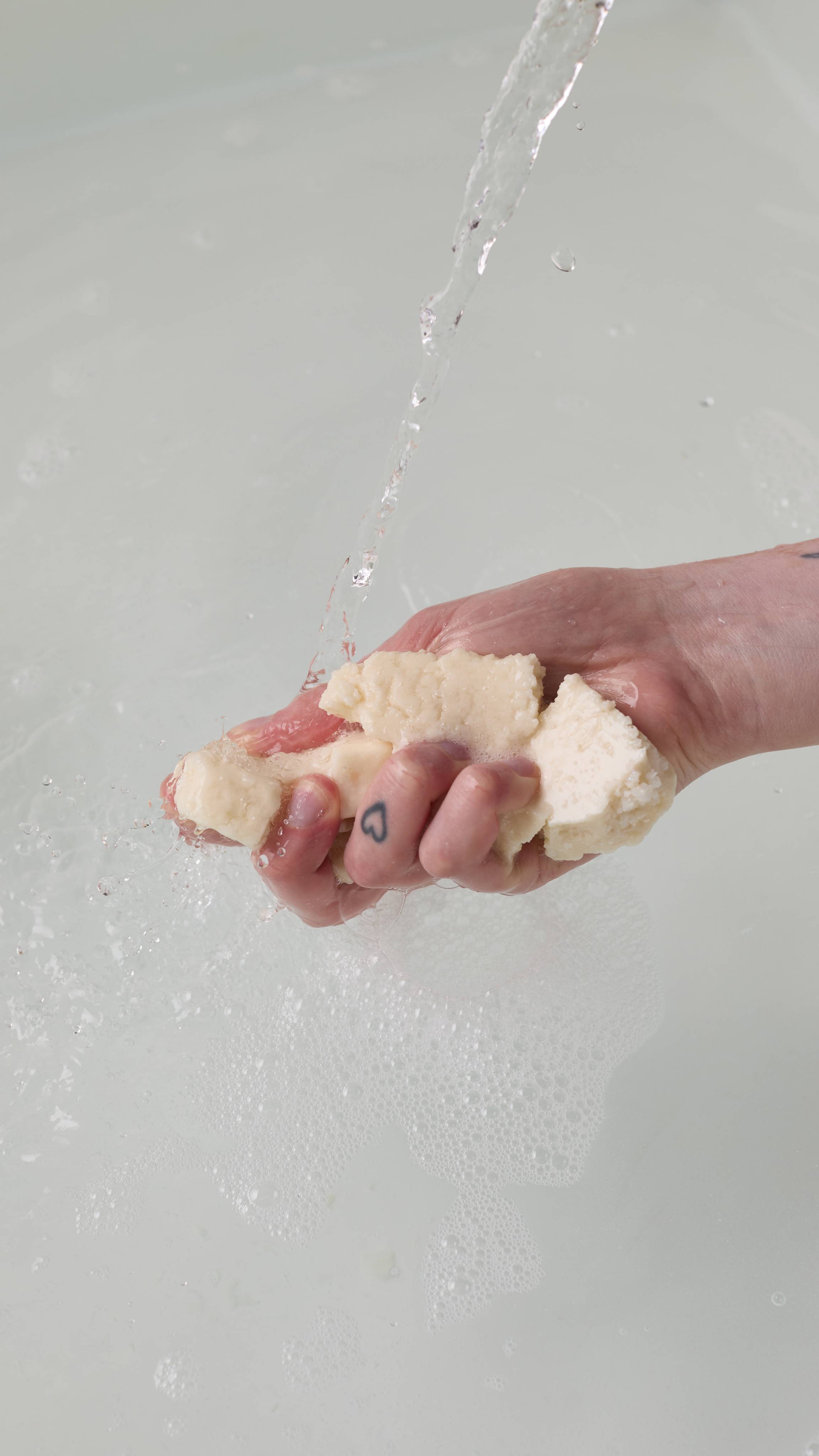 The image shows the model's hand reaching in from the right as it crumbles the creamy, crumbly Santa Moritz bubble bar under running tap water in a white porcelain bath. The water has a delicate layer of bubbles forming.