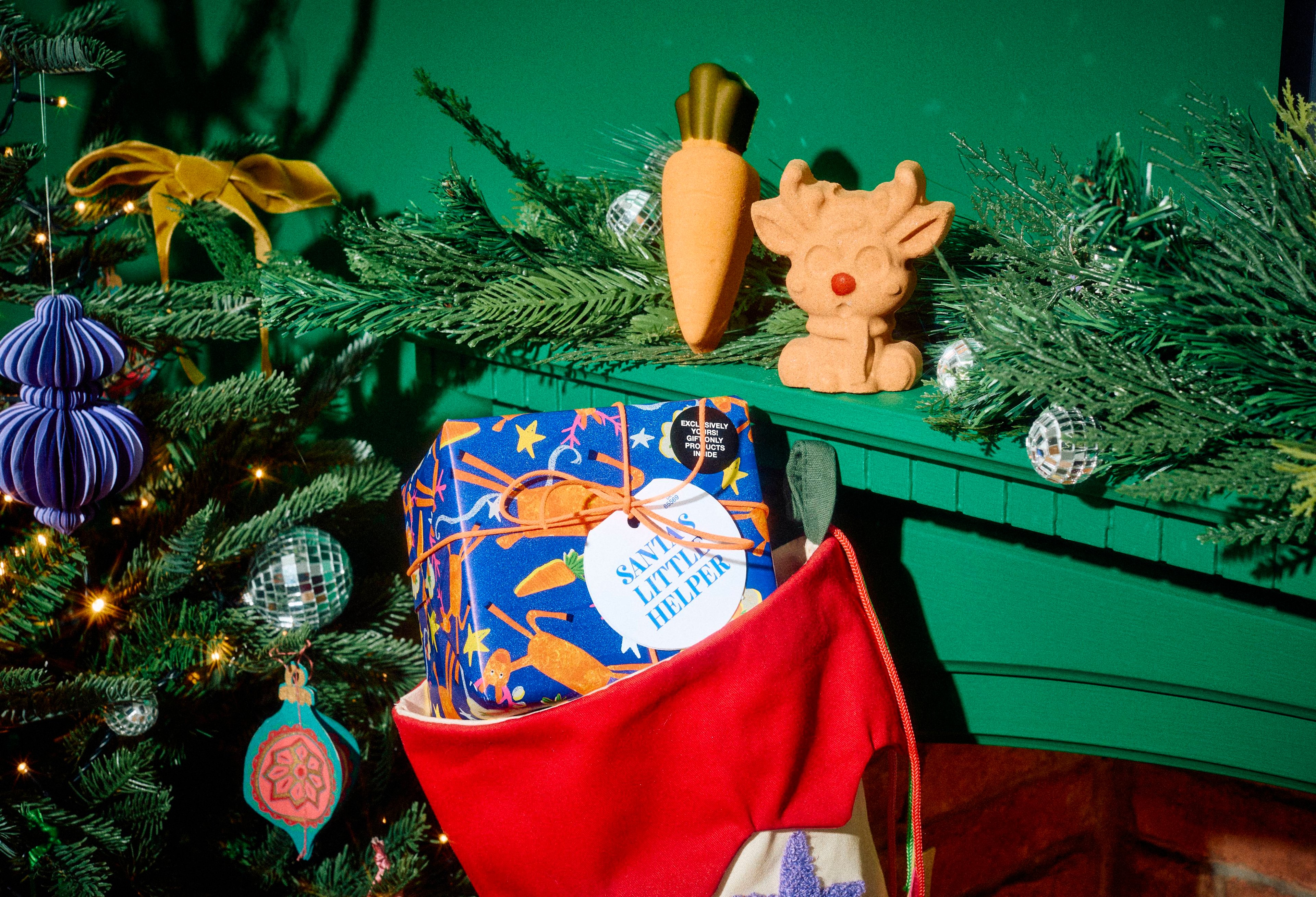 The Lush Santa's Little Helper gift box tucked inside a red stocking, with an orange carrot bath bomb and reindeer bath bomb displayed on a green mantelpiece surrounded by festive greenery.
