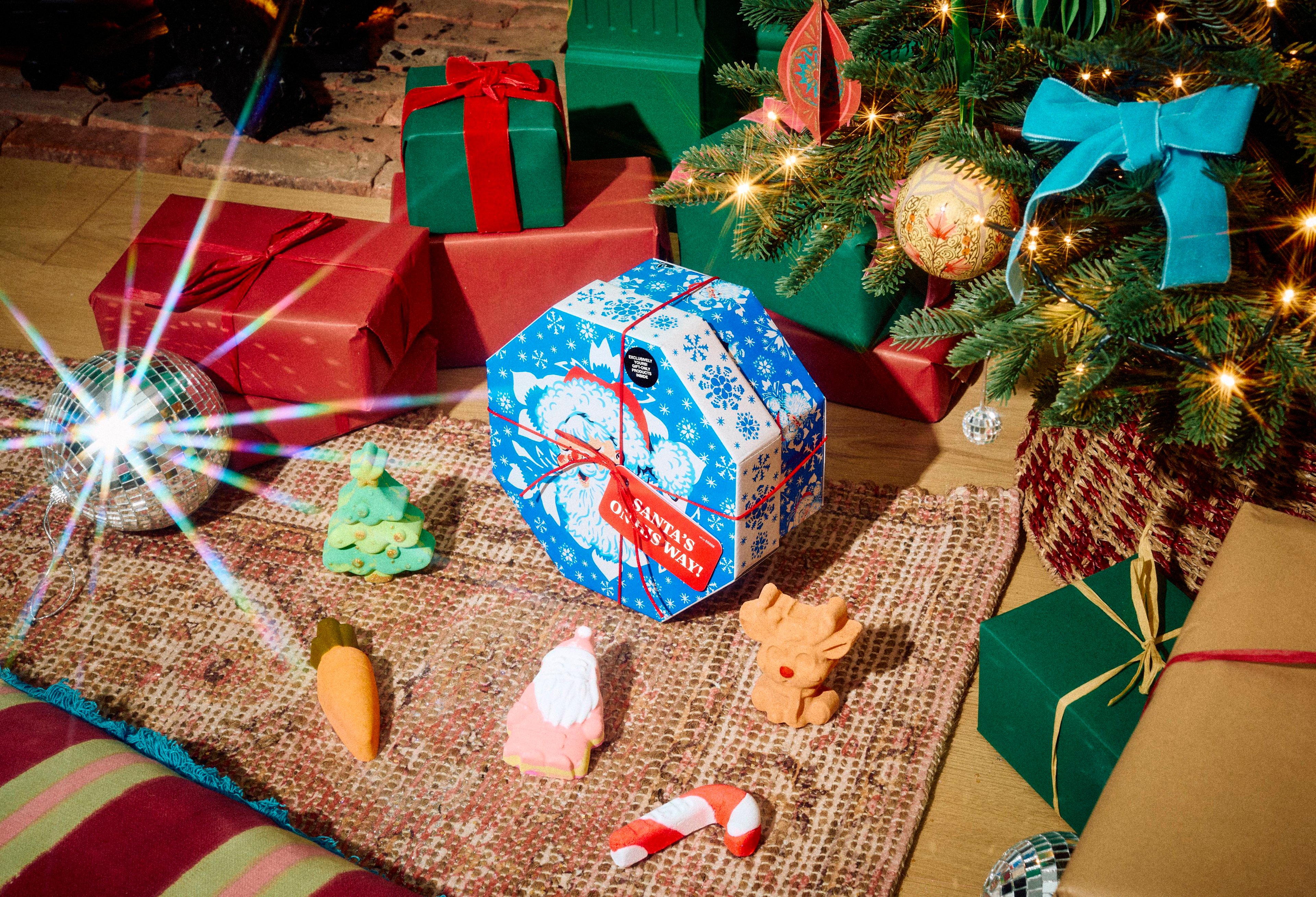 The Lush Santa's On His Way gift set sits on a patterned rug surrounded by festive bath products, including a Christmas tree, reindeer, Father Christmas, candy cane, and carrot bath products, in a room with presents under a Christmas tree.