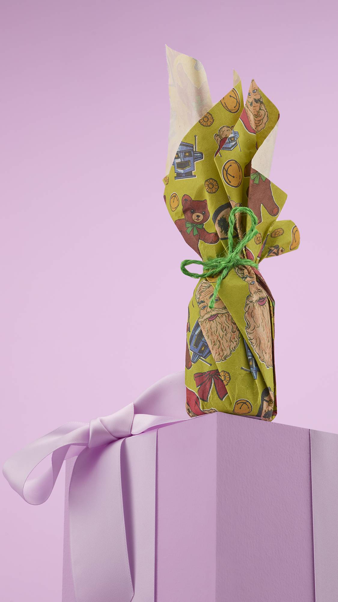 A festive, tall, wrapped gift with yellow and green paper featuring a cartoon Santa Claus and teddy bears. The gift is tied at the top with a green string and is sitting on a light purple, ribbon-wrapped box.