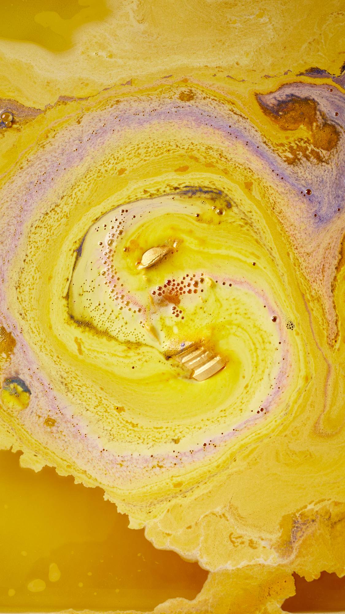 An aerial view of the festive, Lush, Santa's Postbox dissolving in the bath water. There is a thick, velvety, sunshine-yellow foam coming off the product with bursts of contrasting pastel-purple from the hollow centre.