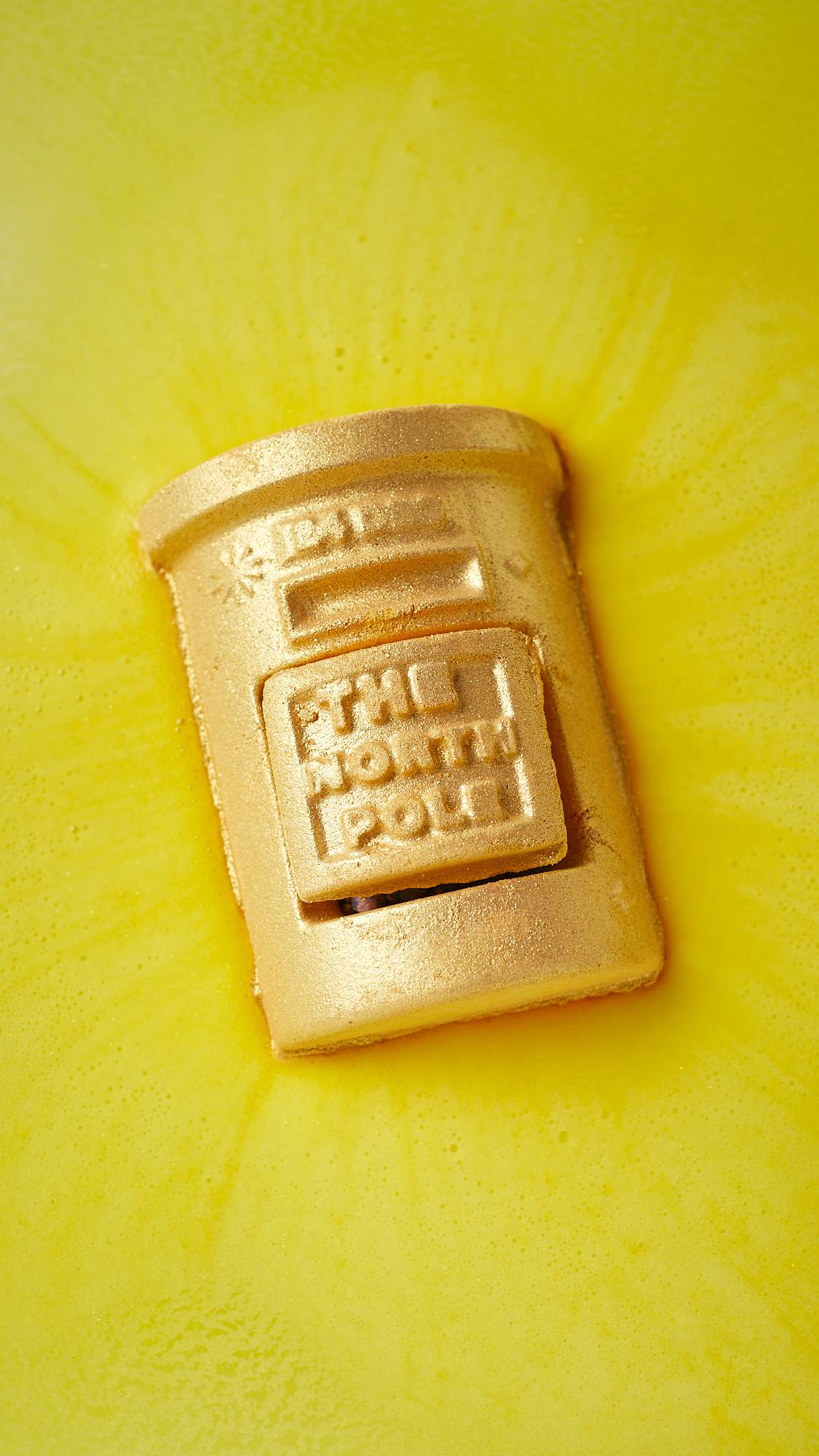 An aerial view of the festive, Lush, Santa's Postbox sitting on the surface of the water as it slowly begins to dissolve. There is a thick, velvety, sunshine-yellow foam coming off the product. 
