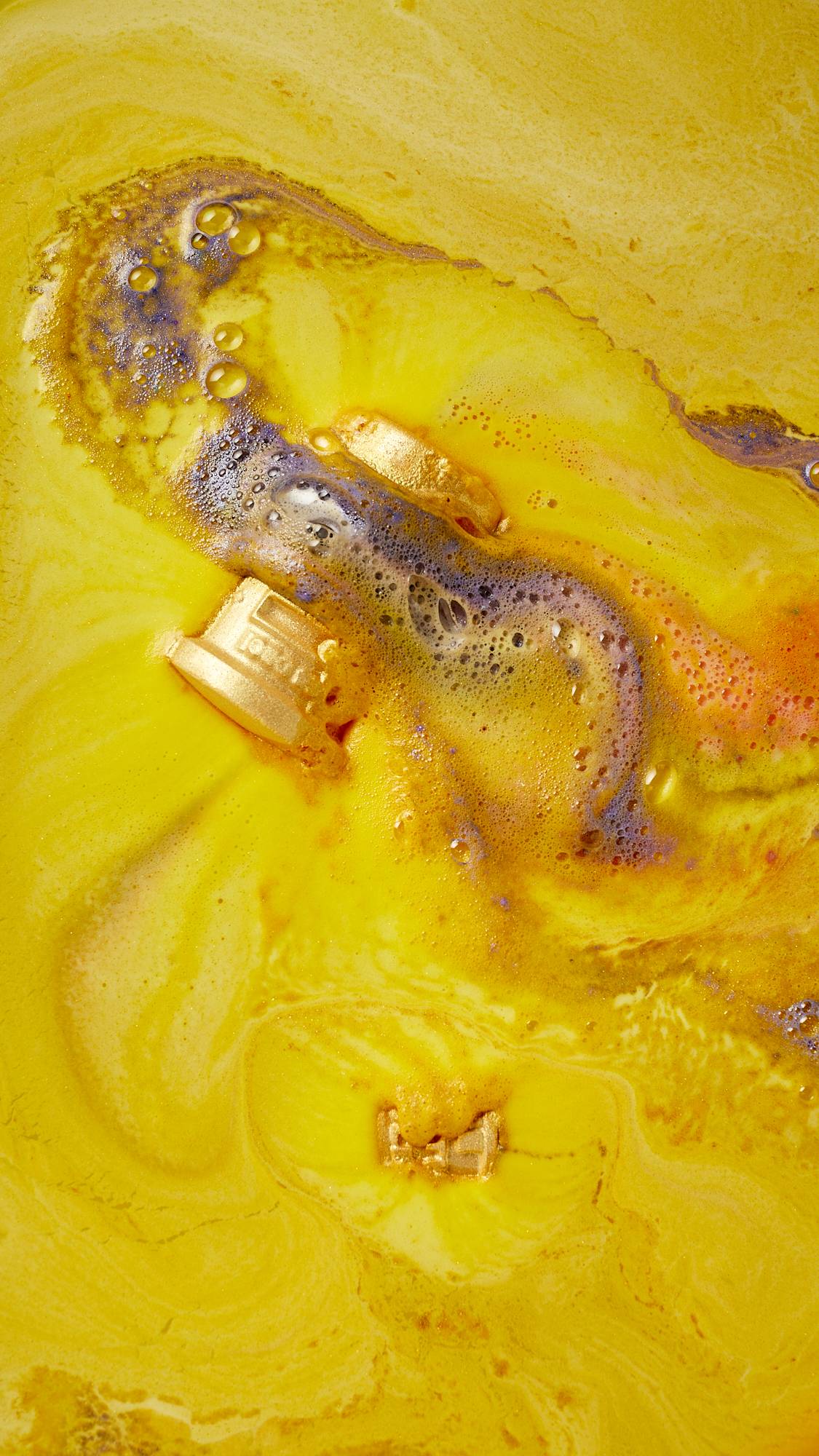 An aerial view of the festive, Lush, Santa's Postbox dissolving in the bath water. There is a thick, velvety, sunshine-yellow foam coming off the product with bursts of contrasting pastel-purple from the hollow centre.