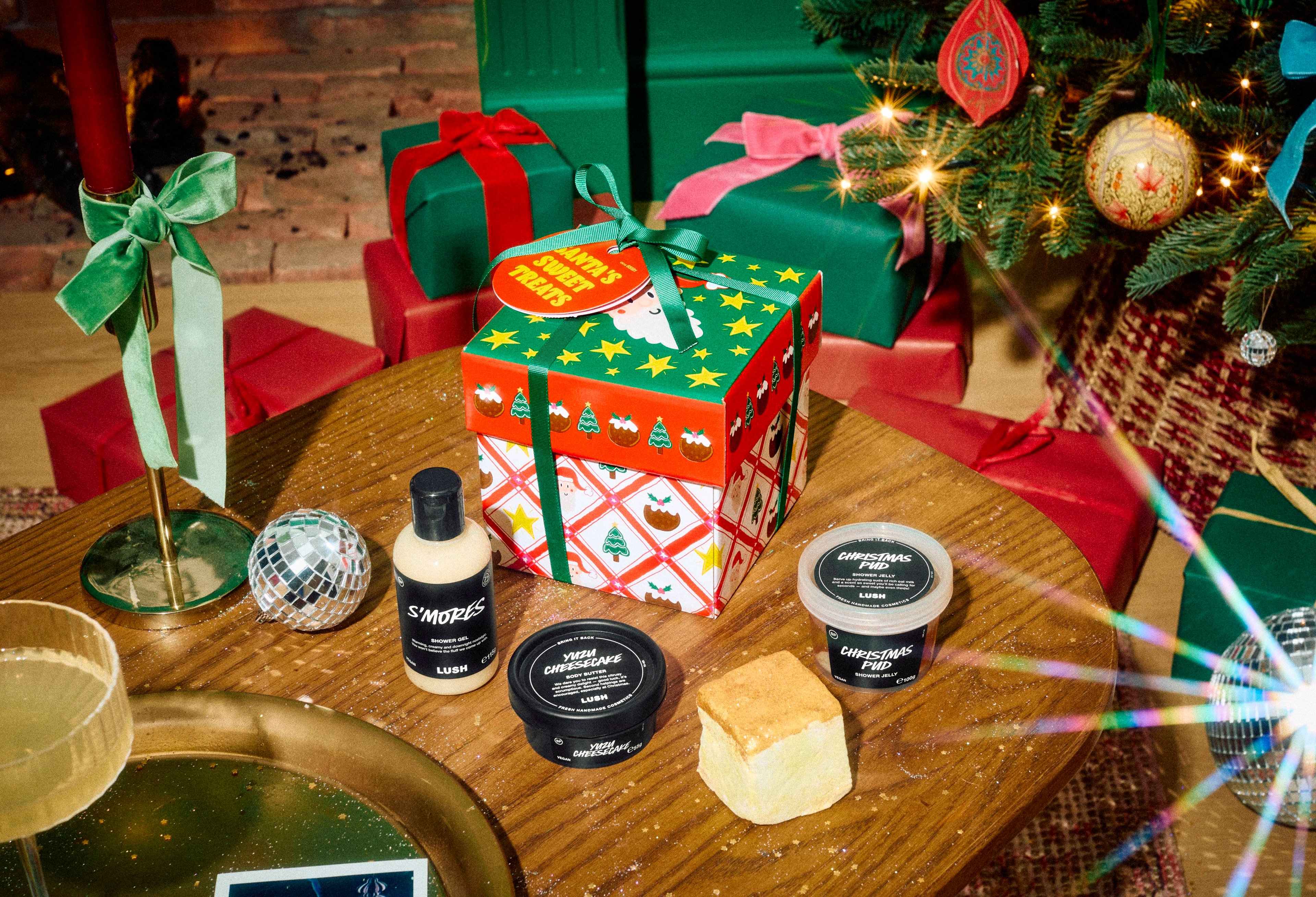The Lush "Santa's Sweet Treats" gift box sits on a wooden table with S'mores shower gel, Yuzu Cheesecake body butter, Christmas Pud shower jelly, and a soap slice, set in a festive room with wrapped presents and a Christmas tree.