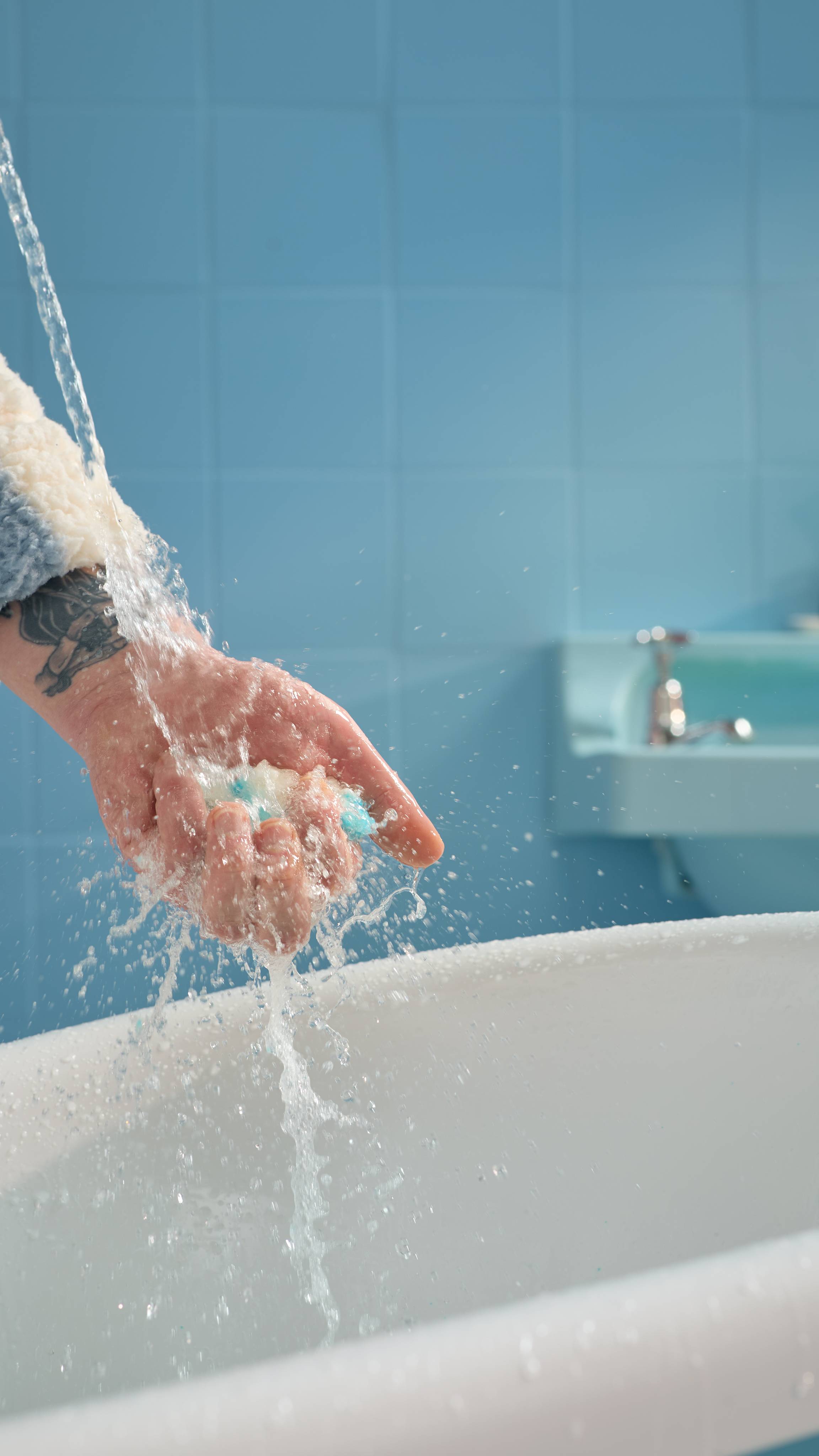The image shows the model's hand over a bath tub holding the Seaside bath bomb as water splashes everywhere in a blue, tiled bathroom. 