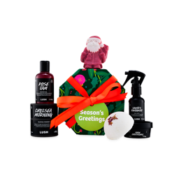 A Lush Season's Greetings gift set tied with an orange ribbon, surrounded by Rose Jam shower gel, Chelsea Morning dusting powder, Santa's Favourite body spray, Yuzu Cheesecake body butter, naked body scrub and Santa-shaped soap.