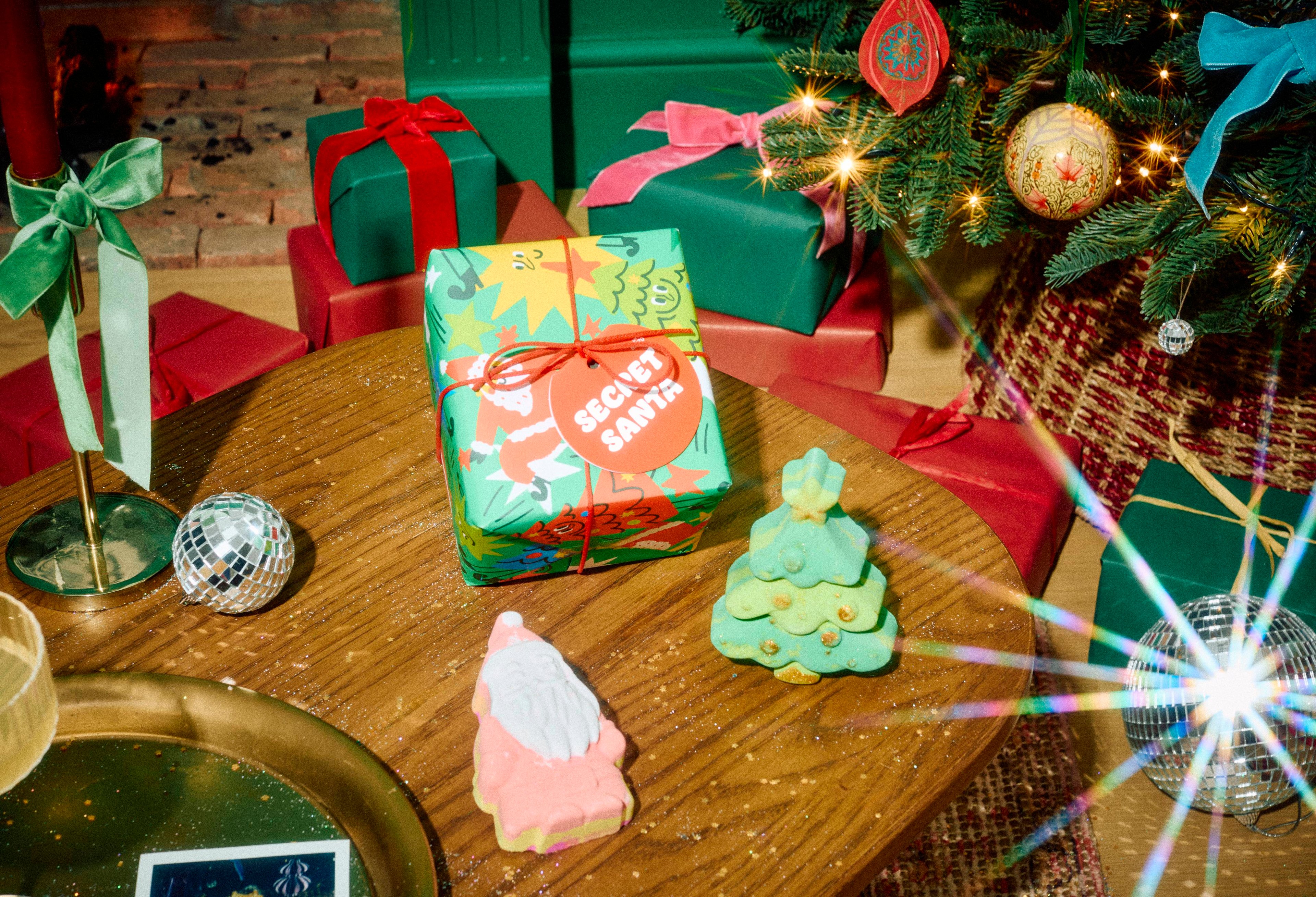 The Lush Secret Santa gift set sits on a wooden table alongside its pink Father Christmas and green Christmas tree bath bombs, set in a festive room with wrapped presents and a decorated Christmas tree.