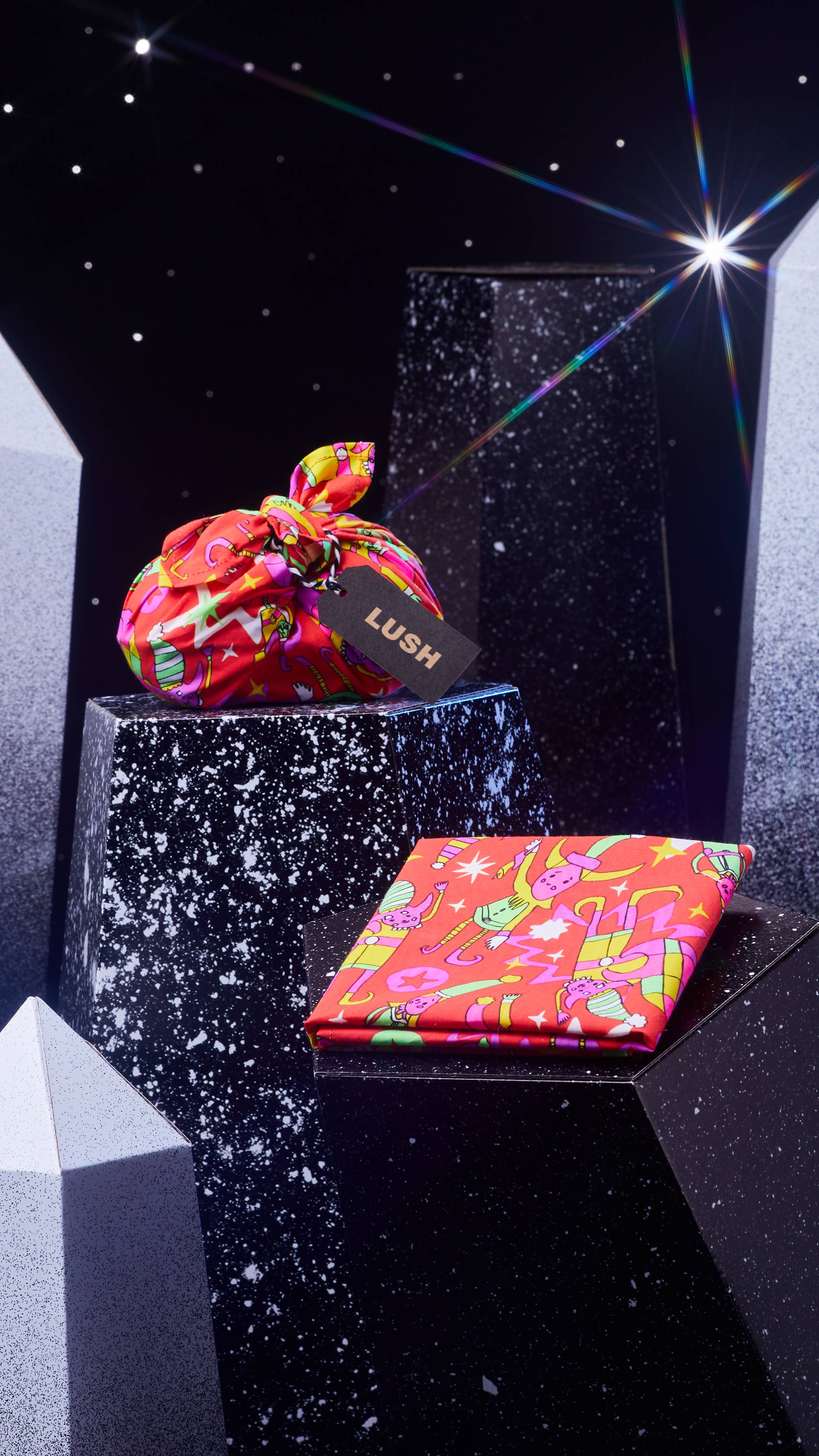 Secret Santa Knot Wrap is shown two ways: folded and wrapping a gift on a dark chromatic platform among geometric shapes.