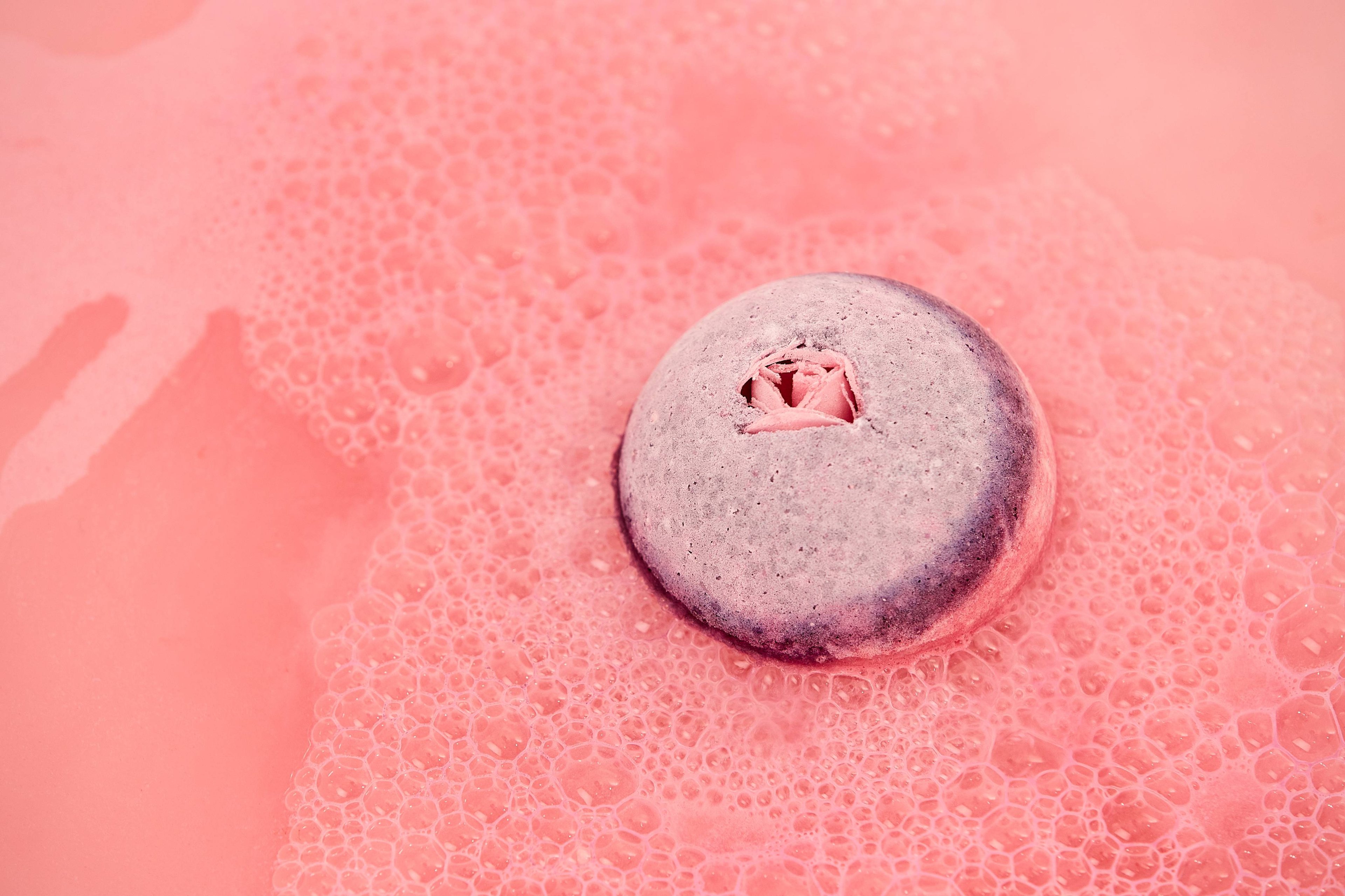 The lilac top and pink rose bud of Sex Bomb bath bomb sinks in pink frothy water.