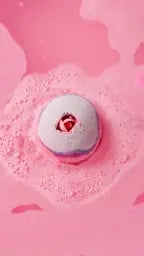 The Sex Bomb bath bomb fizzes in vibrant pink water, creating soft foam. The round purple and pink bomb floats in the center, showcasing its signature rosebud detail.