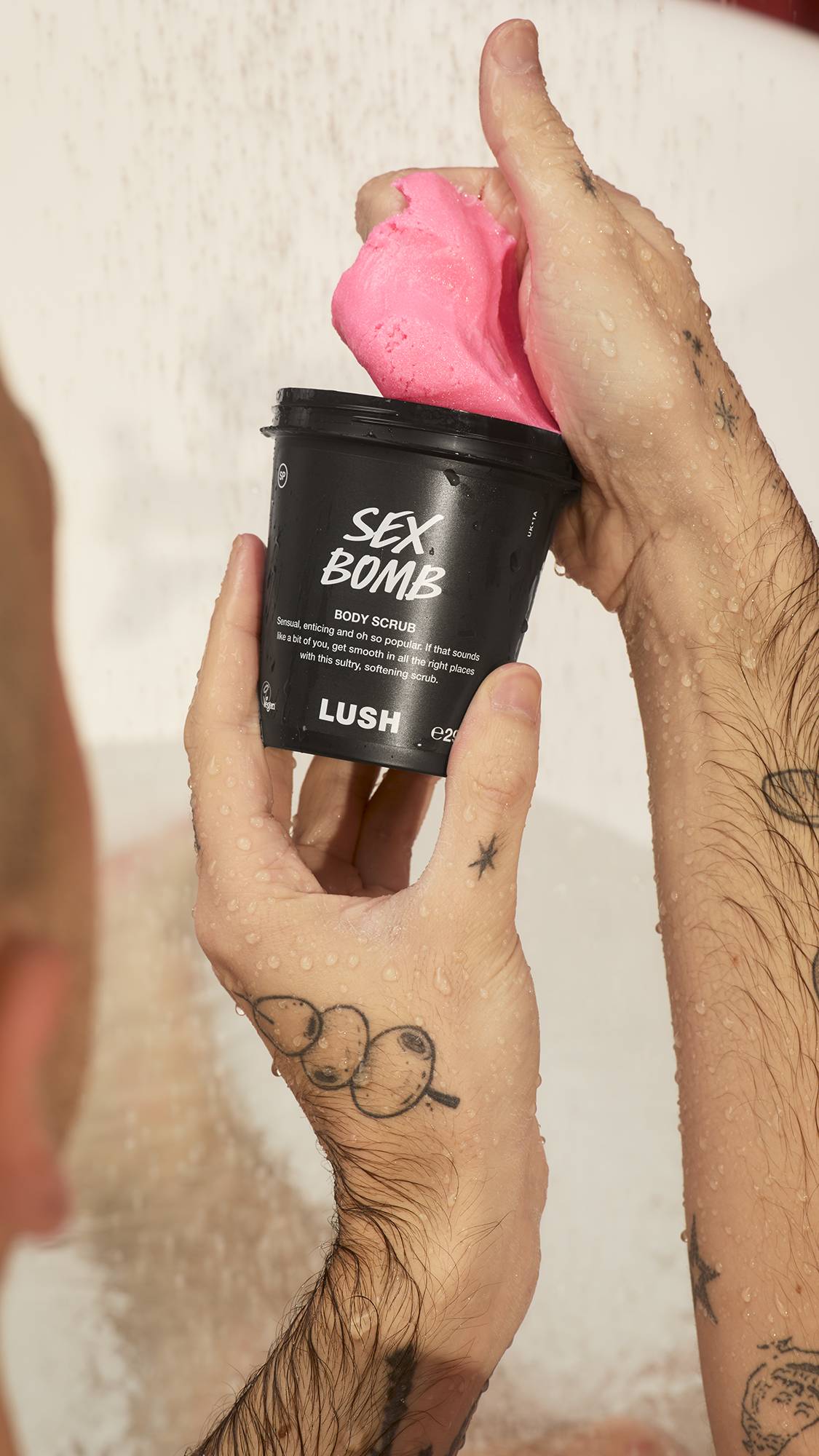 Wet, tattooed hands hold a black pot of Sex Bomb Body Scrub, scooping out a large mound of bright pink product against a light, wet background.