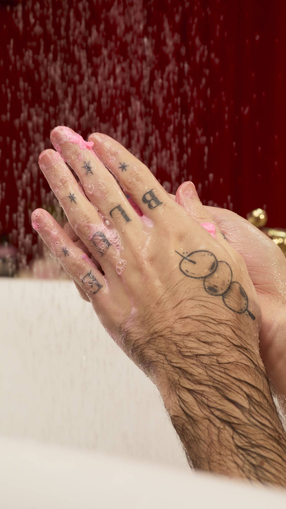 Tattooed hands rub together, lathering the pink Sex Bomb Body Scrub into suds against a wet, dark red background.