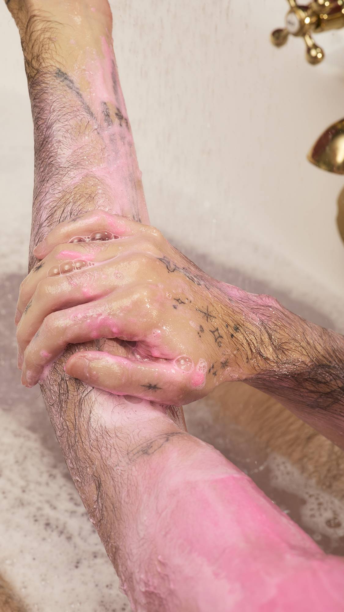 Tattooed hands massage the creamy pink lather of Sex Bomb Body Scrub onto a forearm, with a gold tap and bath water visible in the background.