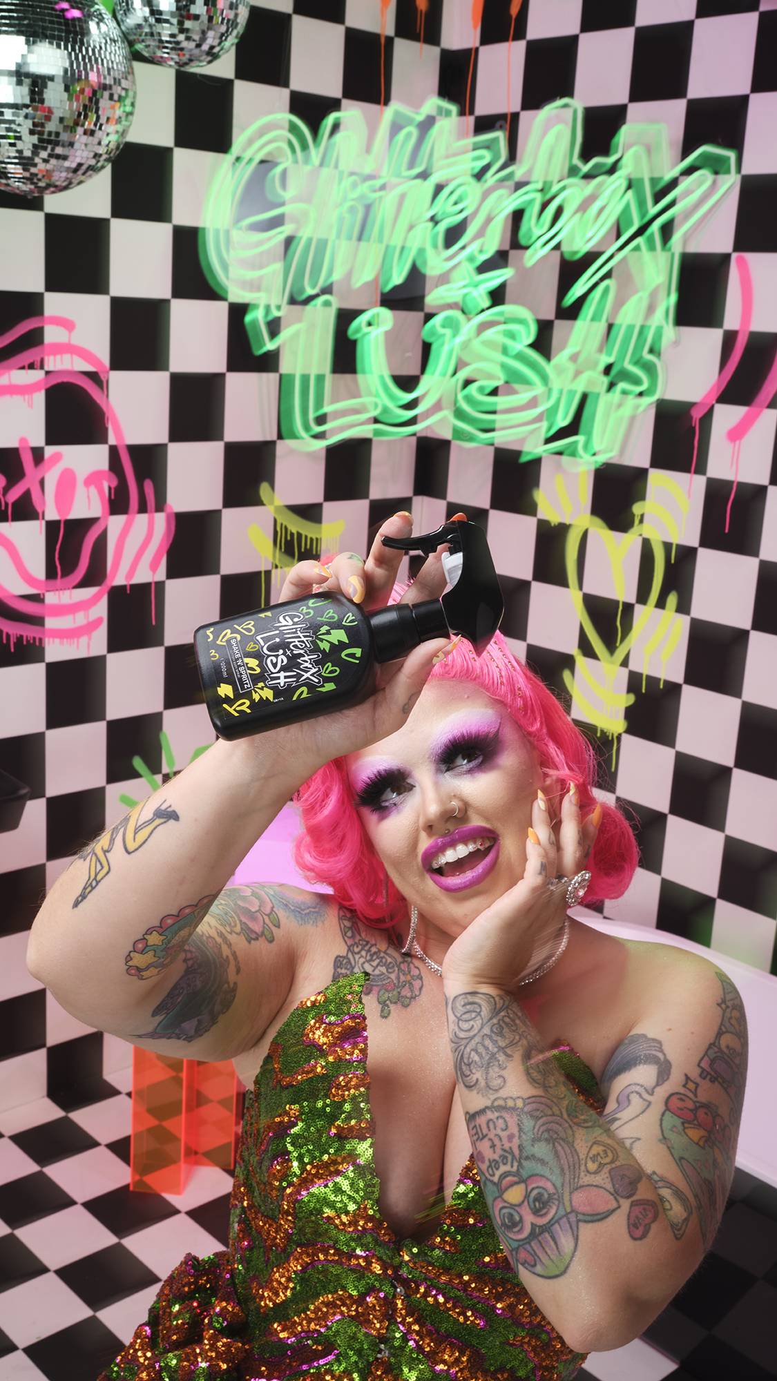 Model has pink hair and a sequin dress holding the spray bottle. They are in a checkerboard room with neon signs and graffiti.