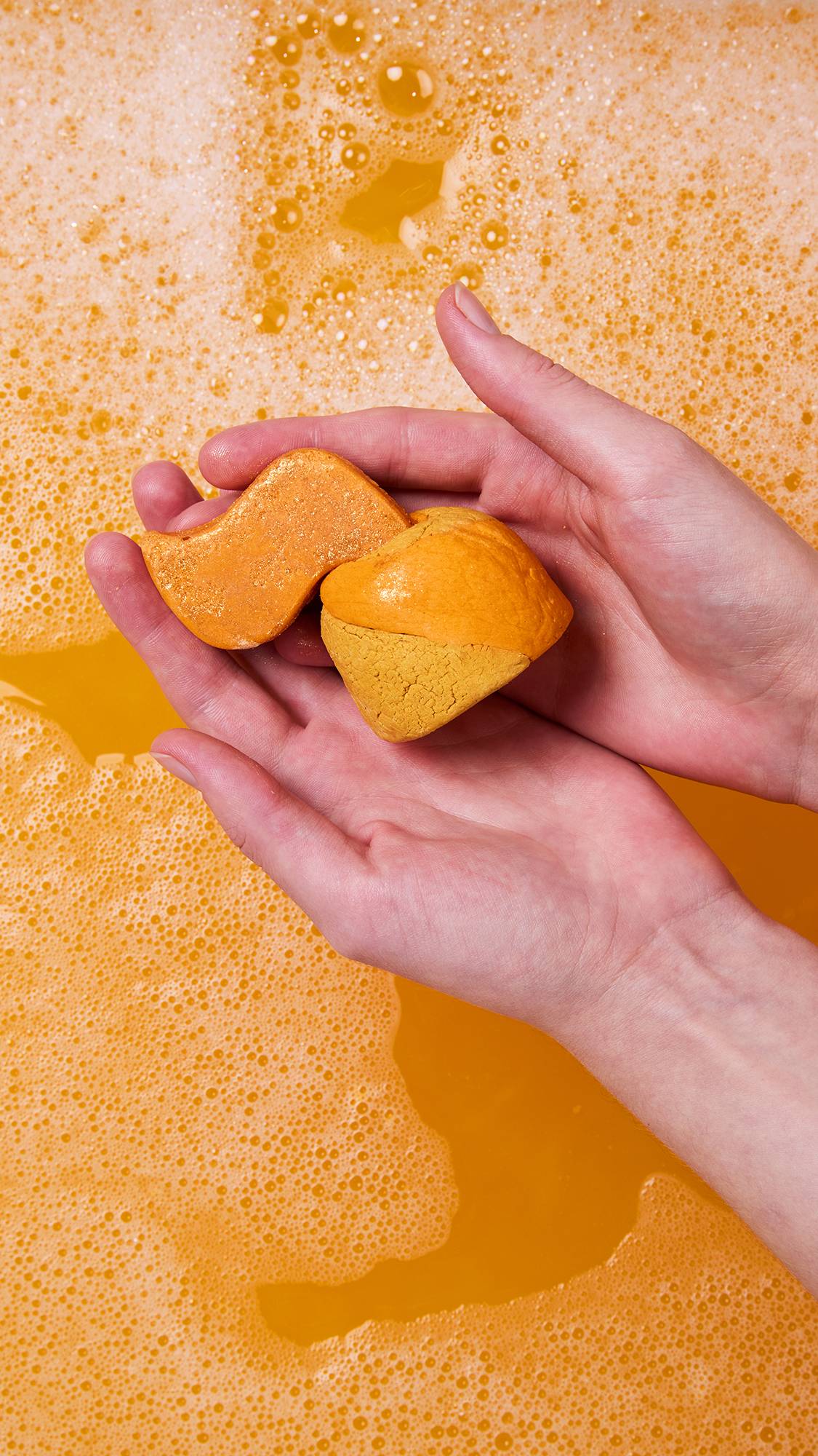 Two hands hold the Shamash Bubble bar over a bubbly, bright orange bath. The bar is broken in two: the rounded, peach/tan bubble bar base and the separate, flame-shaped caramel-amber bath melt component.