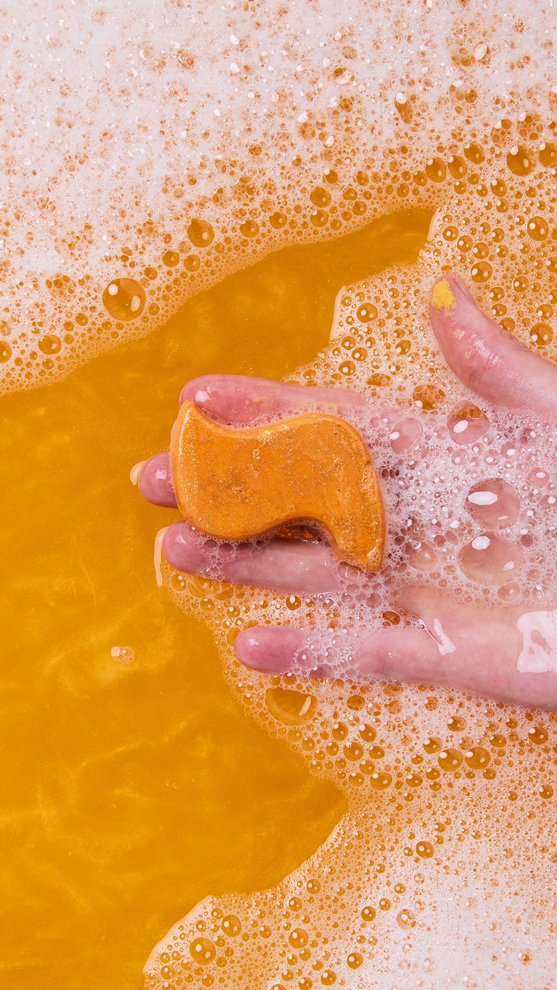 A hand holds the flame-shaped bath melt from the Shamash Bubble bar just above the surface of the bubbly, bright orange bath water. The hand is covered in white foam from the bubbles.