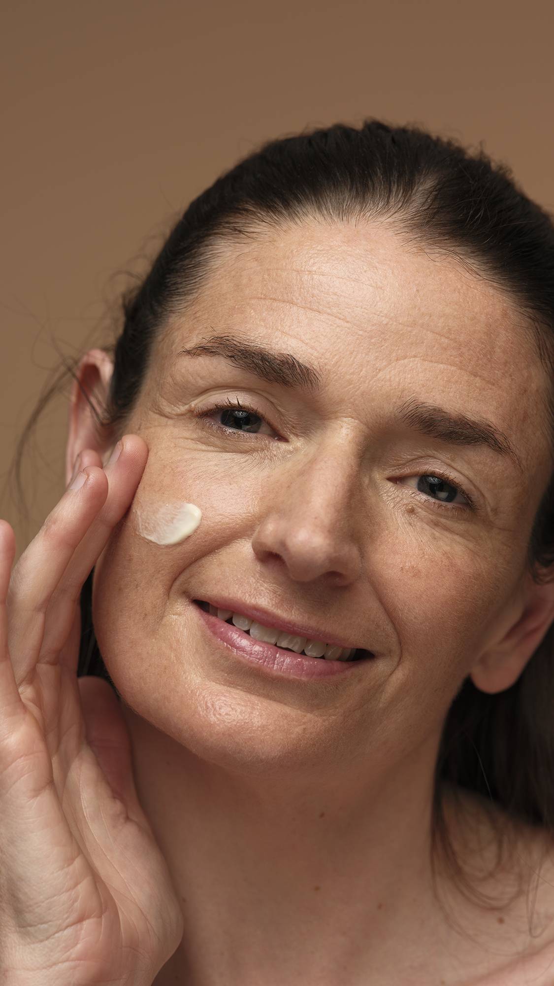 A close-up of the model's face as they gently smooth the Skin's Shangri La self-preserving moisturiser over one of their cheeks. They are on a warm, earthy-brown background.