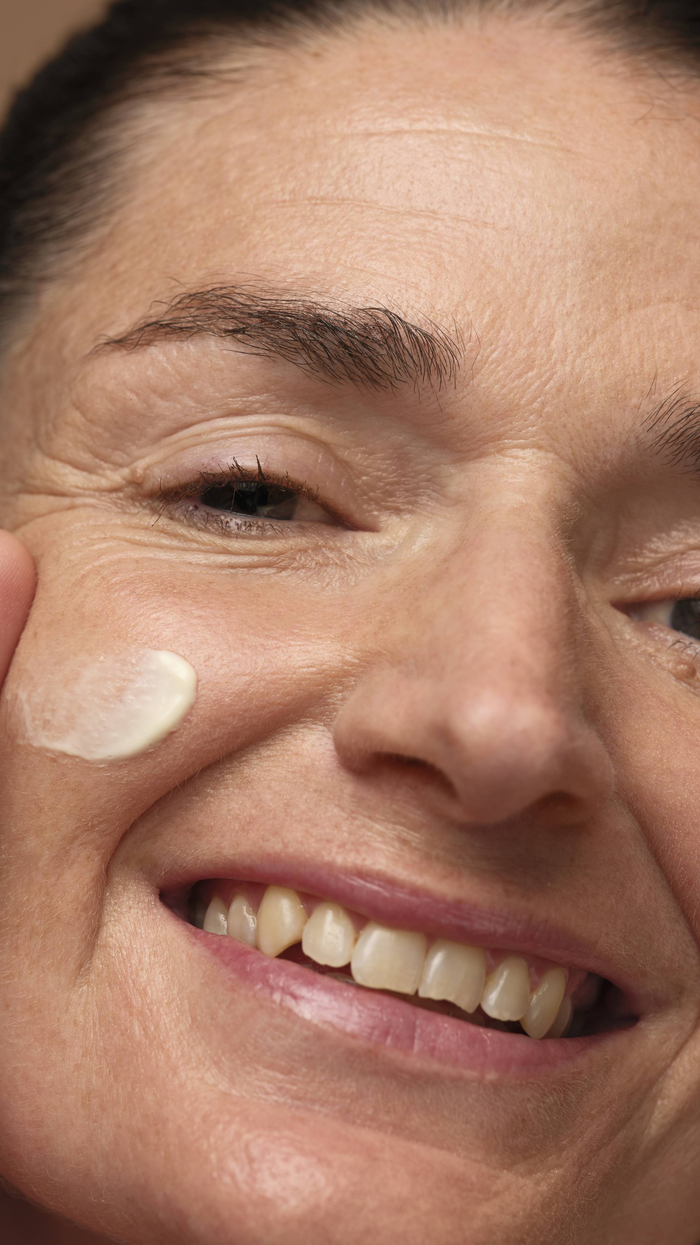 A close-up of half of the model's face as they are smiling while they gently sweep the self-preserving moisturiser over their cheek. 