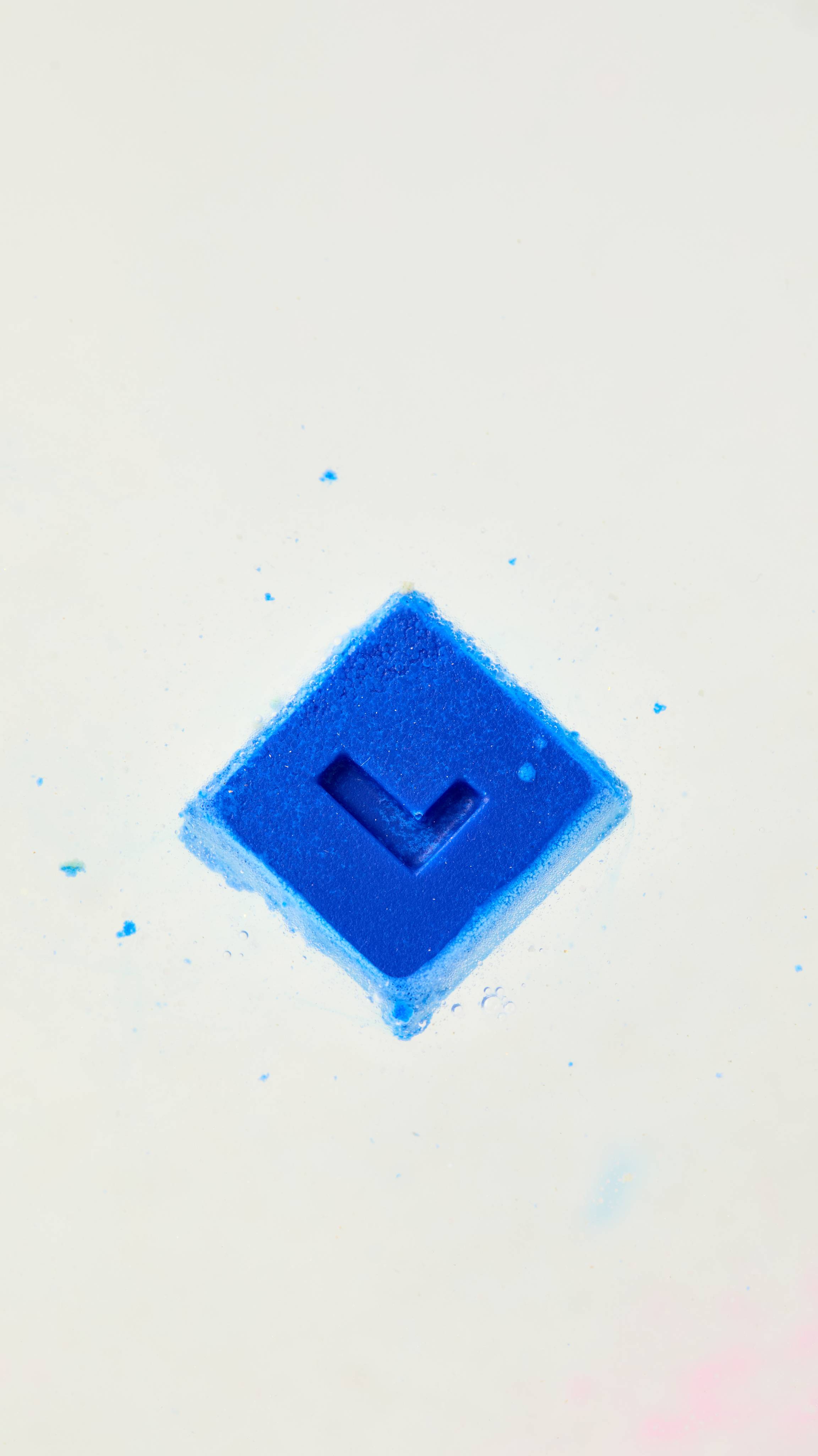 A solid, bright blue, square bath melt floats diagonally in a bath of foamy white water, with a single "L" embossed on its surface and small blue specks visible around it.