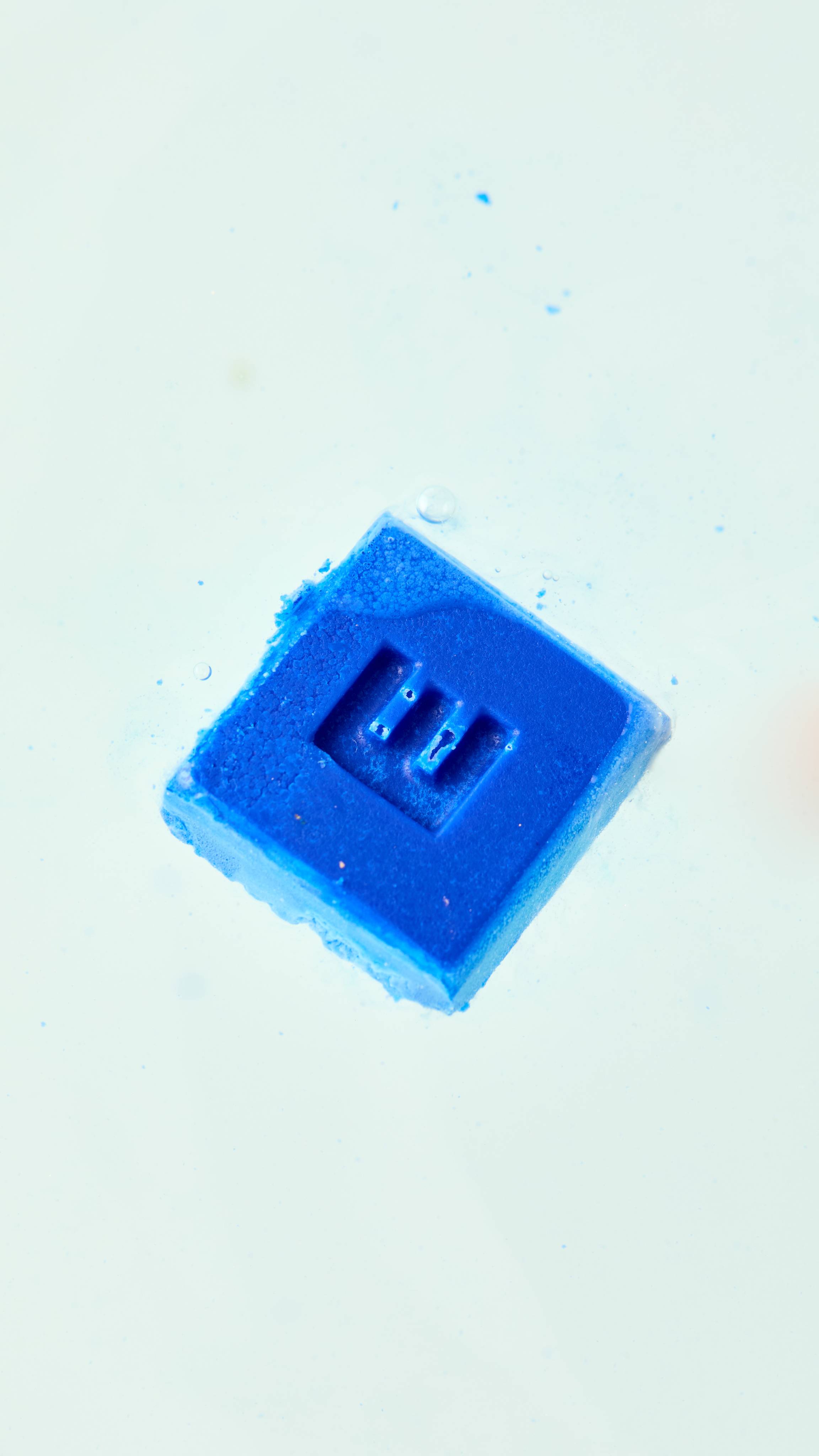 A solid, bright blue, square bath melt floats diagonally in a bath of foamy white water, with a single "E" embossed on its surface and small blue specks visible around it.