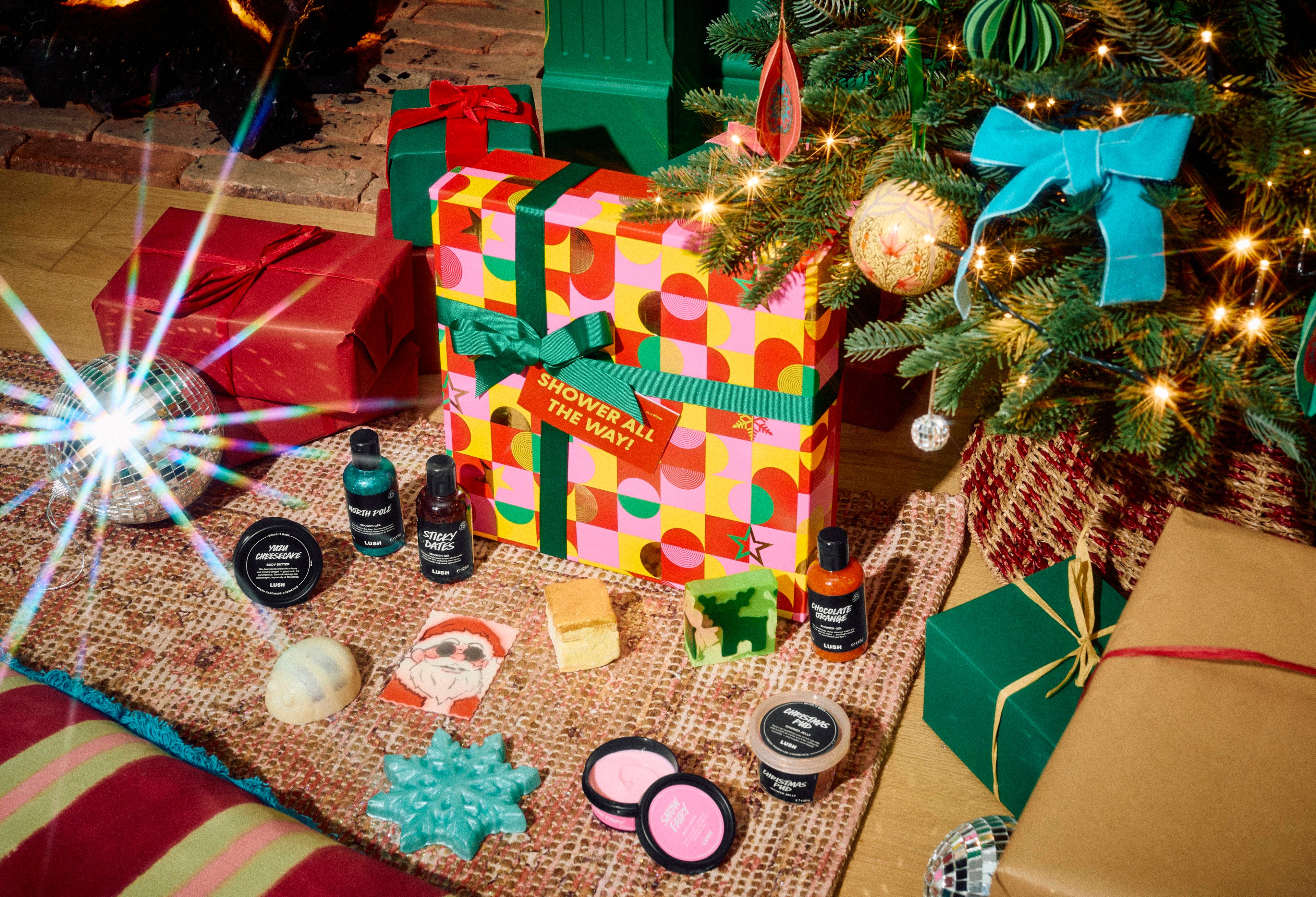 The Lush Shower All The Way! gift box sits on a patterned rug surrounded by 12 Lush products, including shower gels, body scrubs, soaps, and bath products, under a decorated Christmas tree with wrapped presents nearby.