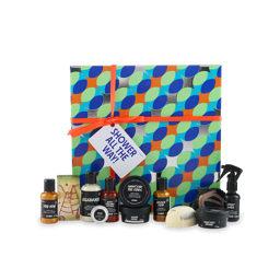 Shower All The Way! A square gift box with a blue, green and orange circle pattern with gold details, tied with an orange ribbon and gift tag. Twelve Lush shower and body care products sit in front of the box.