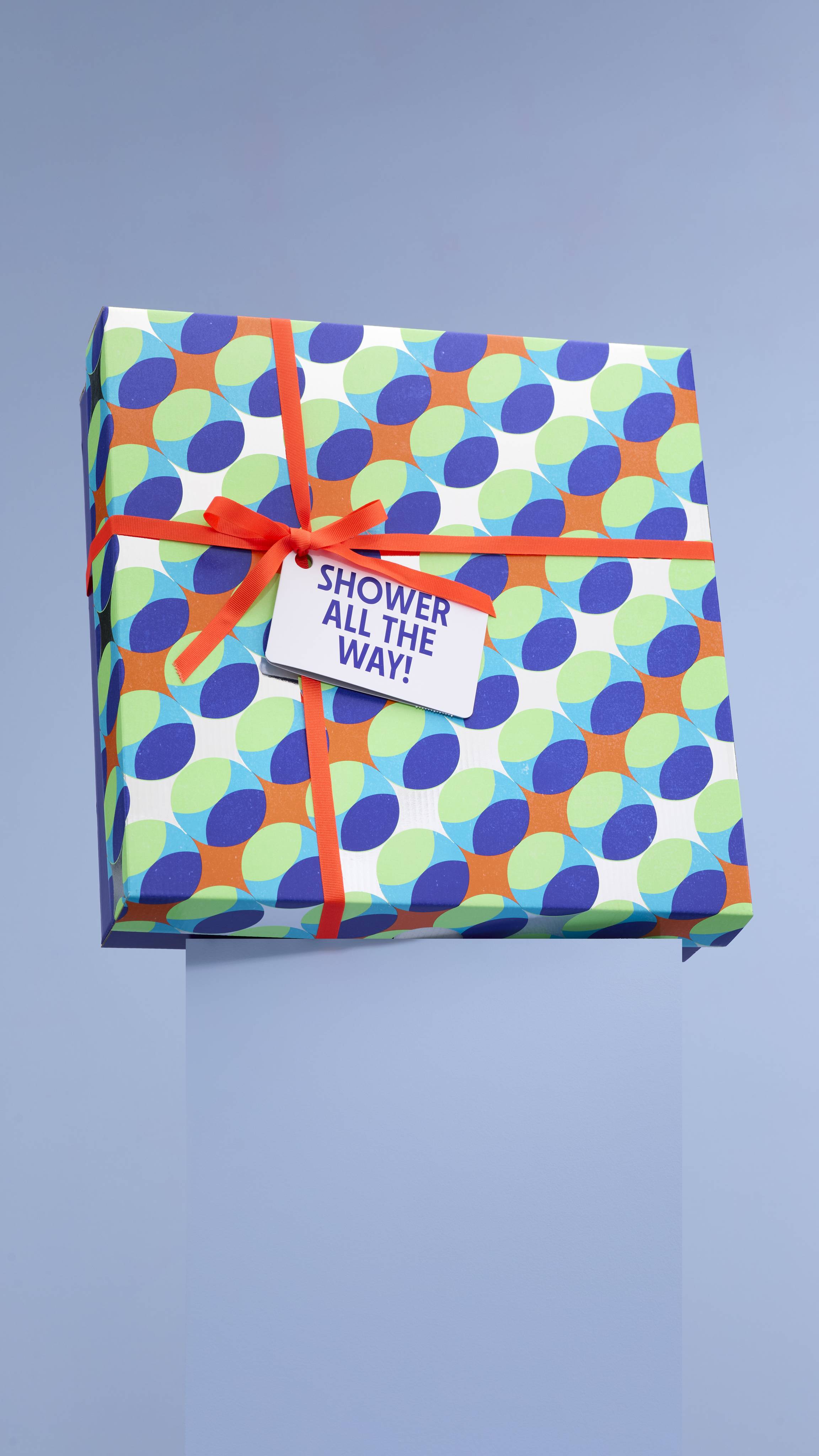 A large, colorful Lush "Shower All the Way" gift box with a geometric pattern of blue, green, and orange shapes. The box is tied with a red ribbon and a tag with the gift set's name.