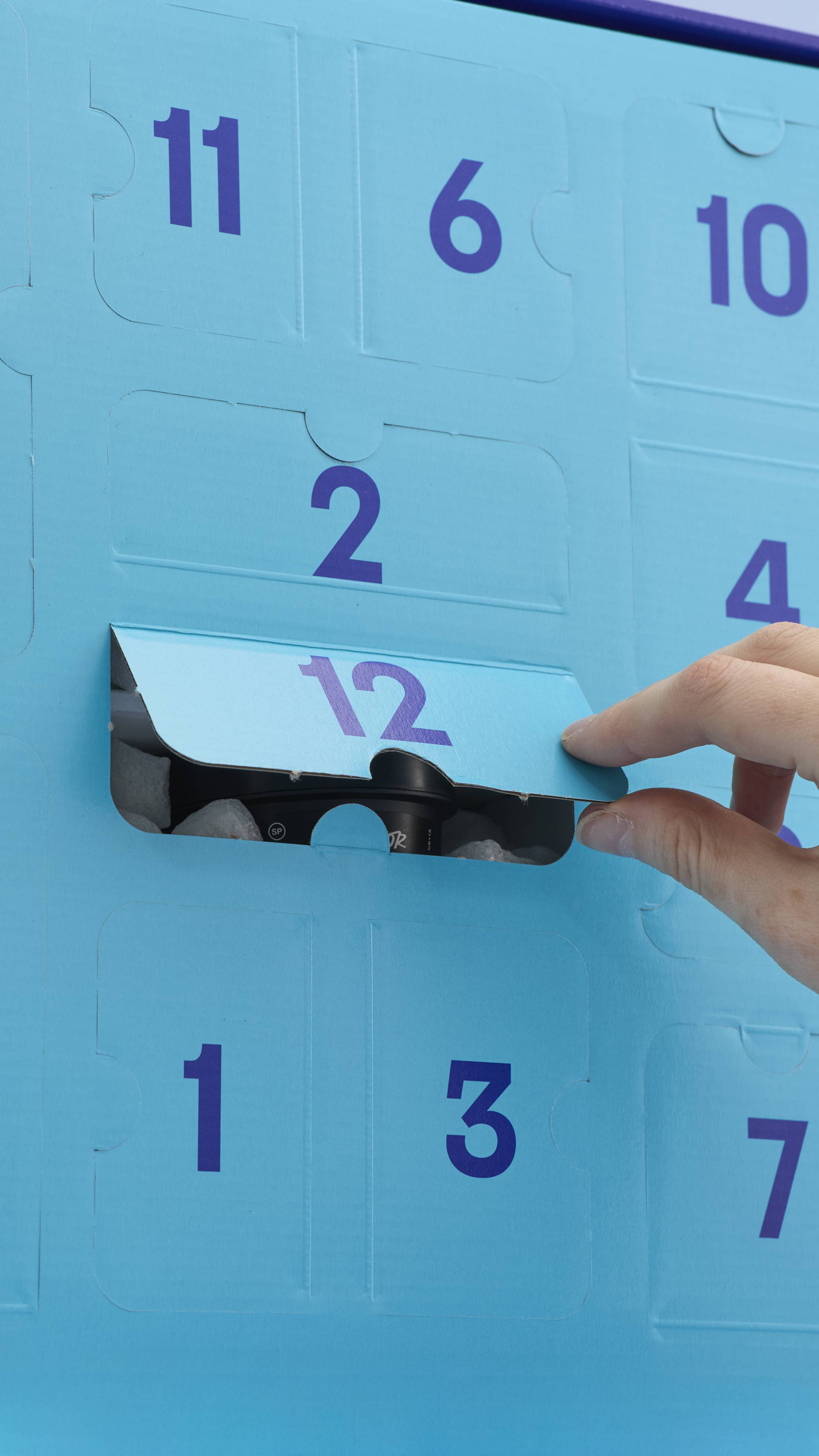 A hand opens door number 12 on the "Shower All the Way" advent calendar. The box has light blue numbered doors with a dark blue interior, revealing a black Lush product inside.