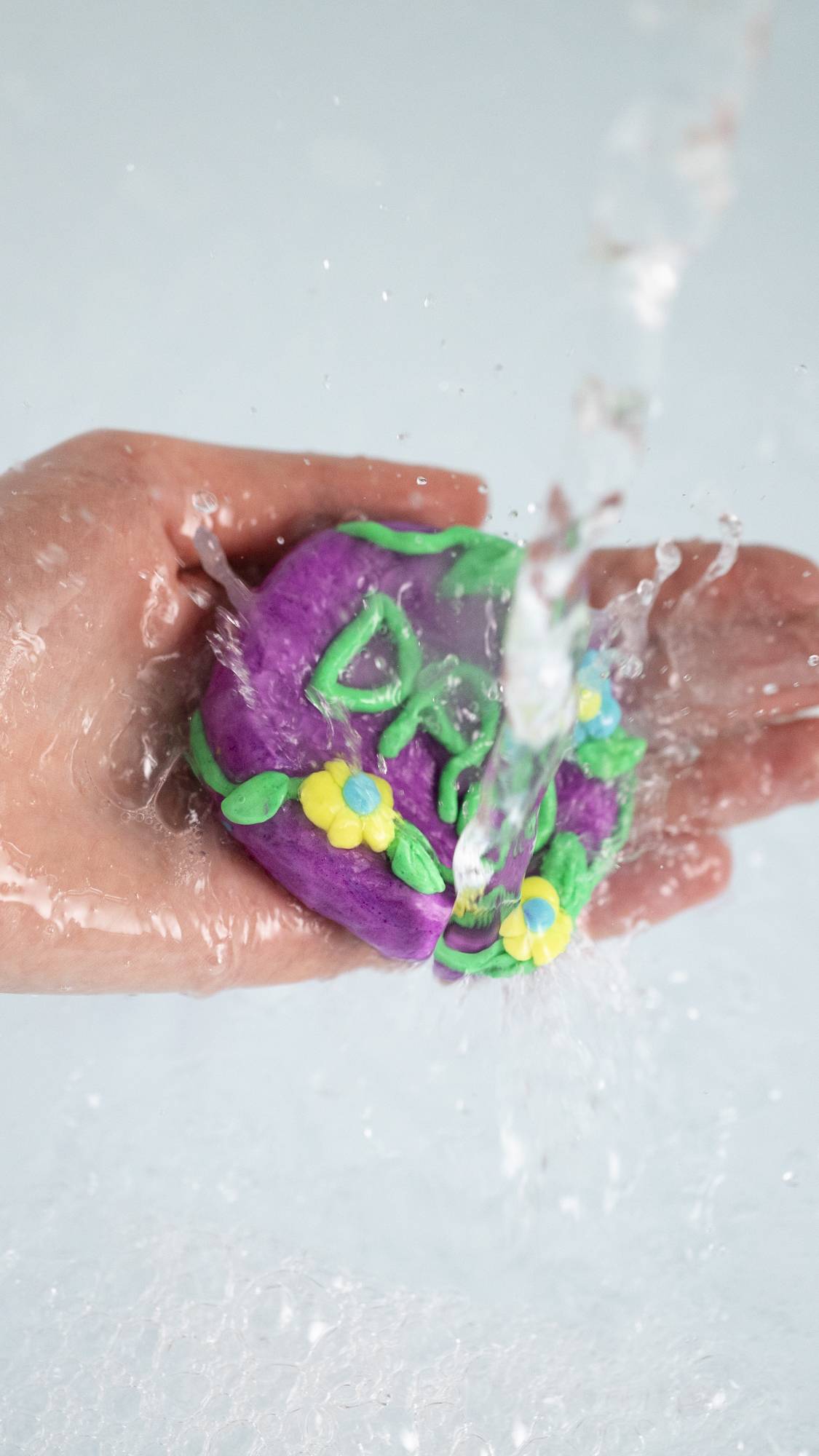 The model is holding the fun product under running water. The product is almost pebble-shaped with flowers and leaves and the word "DAD" in green in the centre.