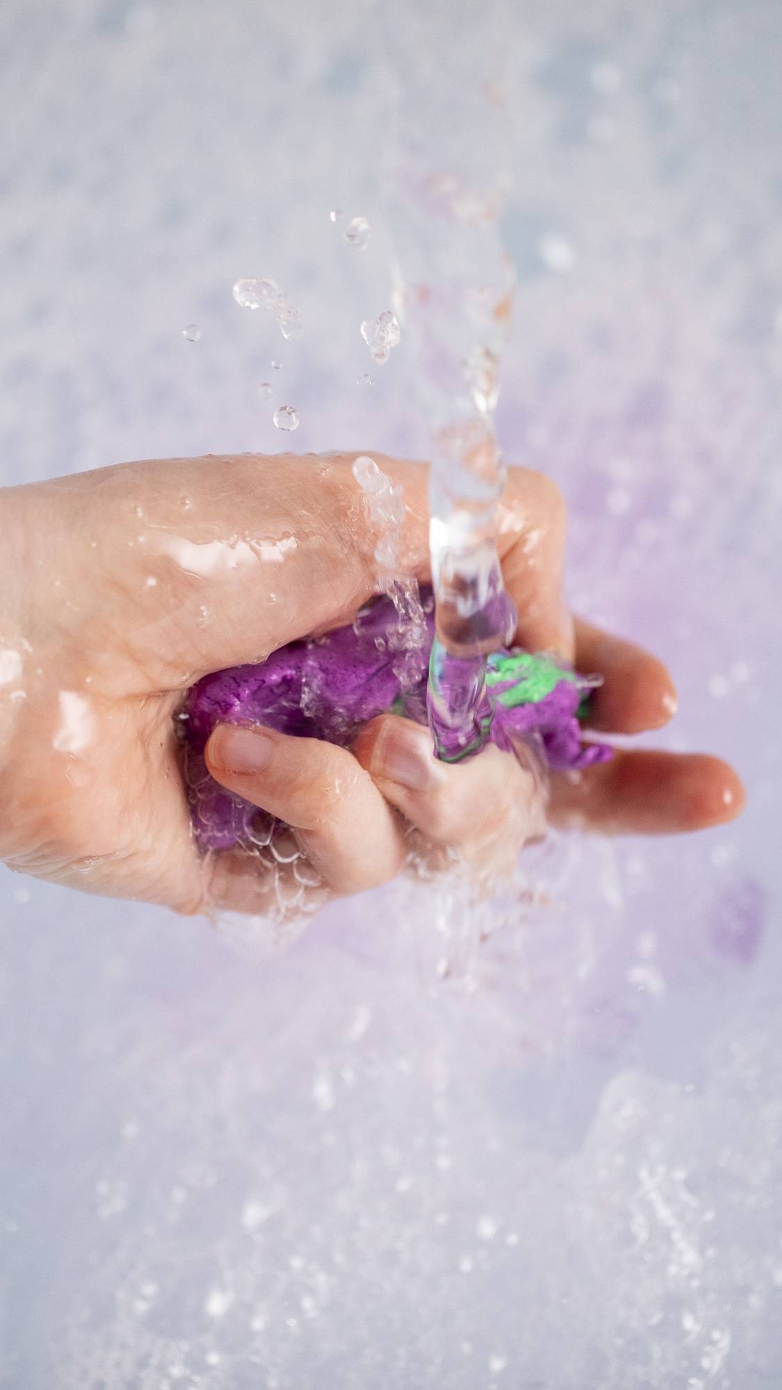 The image shows a close-up of the model's hand as they are squeezing and crumbling the product under running water to create bouncy bubbles below. 