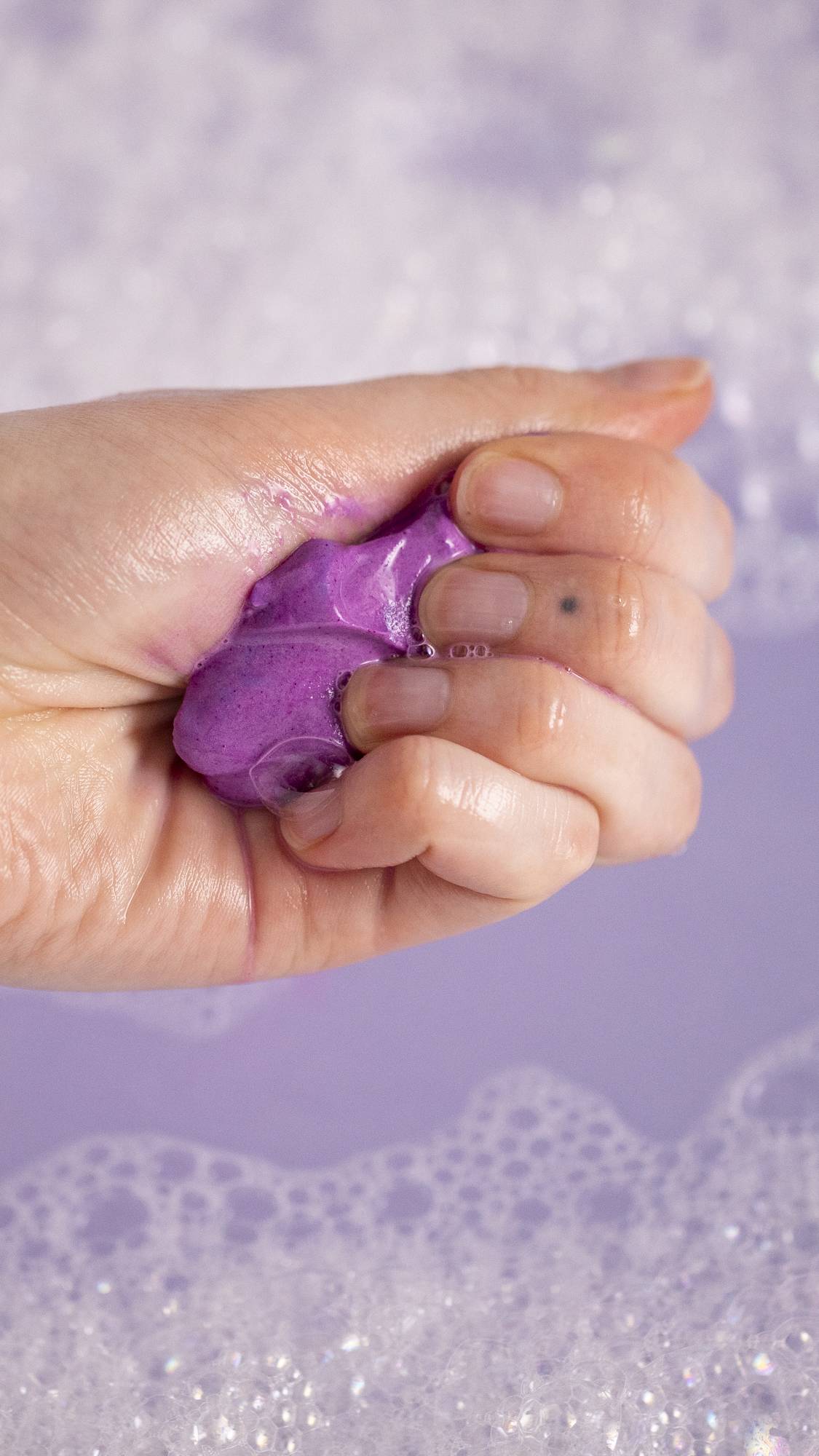 A close-up image of the model's hand as they have squeezed the product into a fun, play-doh-styled lump showing lilac waters and bubbles below. 