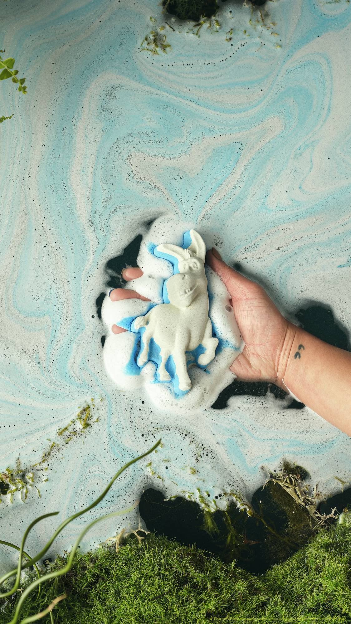The image shows the model's forearm and hand as they hold the Donkey bath bomb just sitting in the ocean-like swirls of blue and white foamy water below. Moss and leaf foliage surround the border. 
