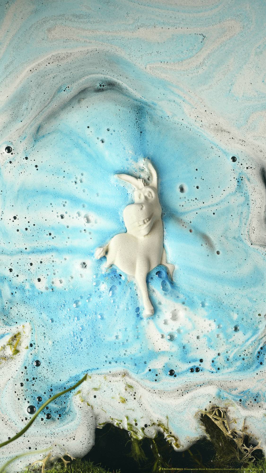 The Donkey bath bomb sits on the surface of the water with the smiling face pointing towards the camera. There are thick, bubbly waves of white and blue foam terminating from the bath bomb. 