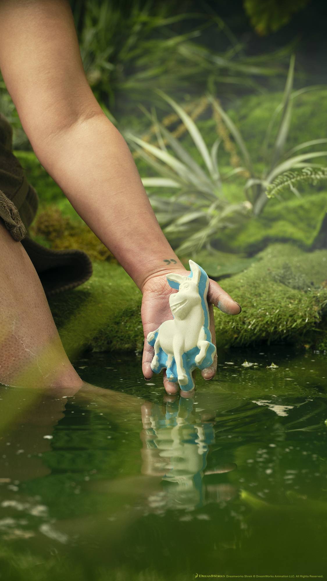A close-up of the model's forearm and hand as they are holding the Donkey bath bob just above the surface of the pond water surrounded by moss and foliage. 