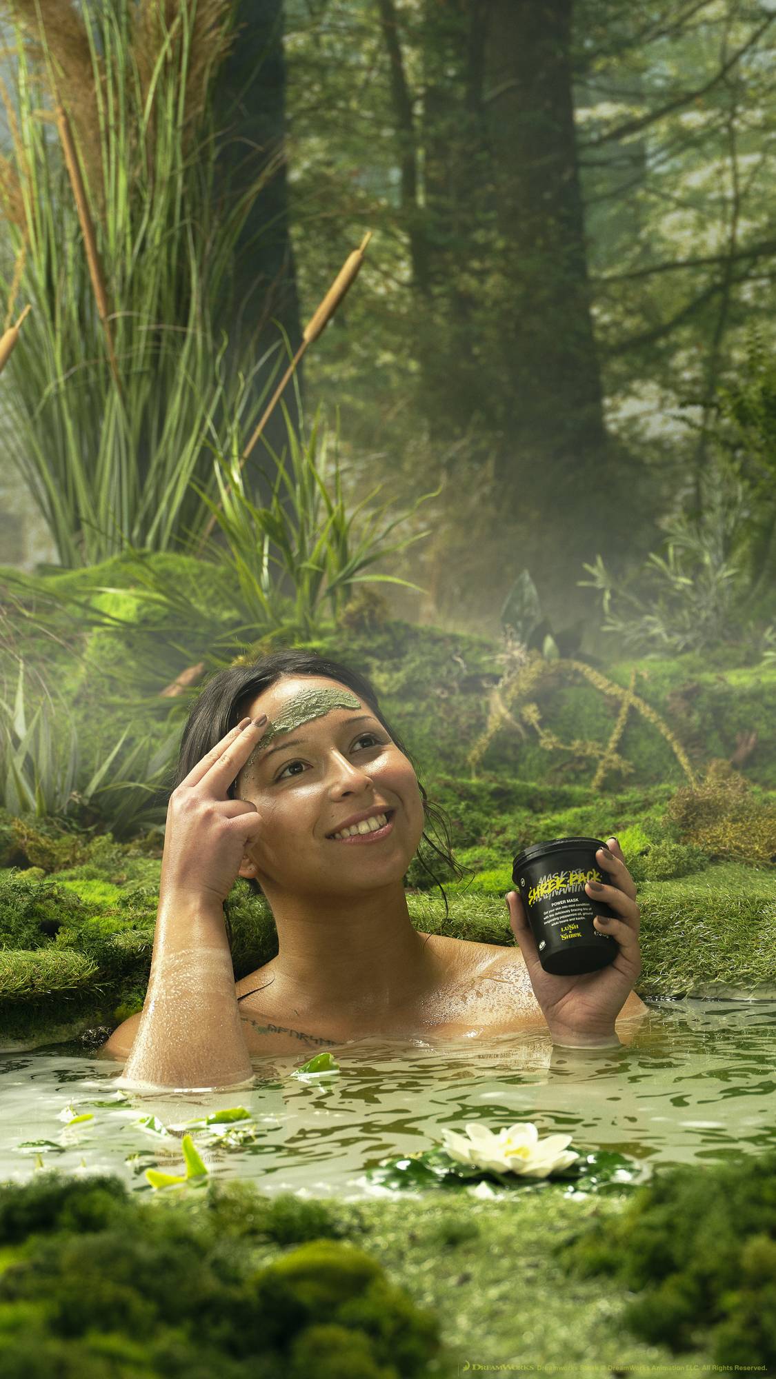 The model is sat in a mossy swamp environment in a spring deep enough to sit in. They are gently applying the Shrek Pack face mask with one hand while holding the product pot with the other. 