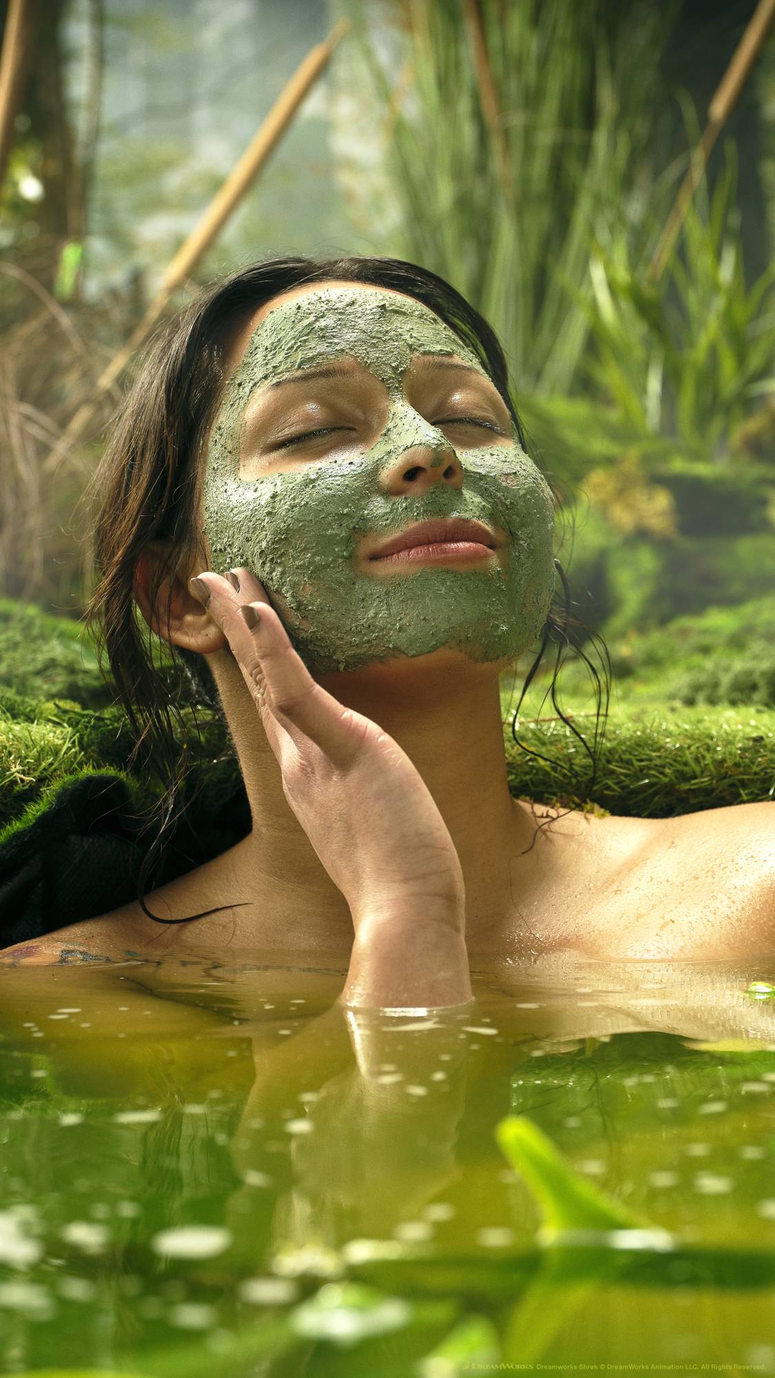 A close-up of the model's face as their eyes are closed and they are leaned back on a mossy knoll with the Shrek Pack face mask covering their face.