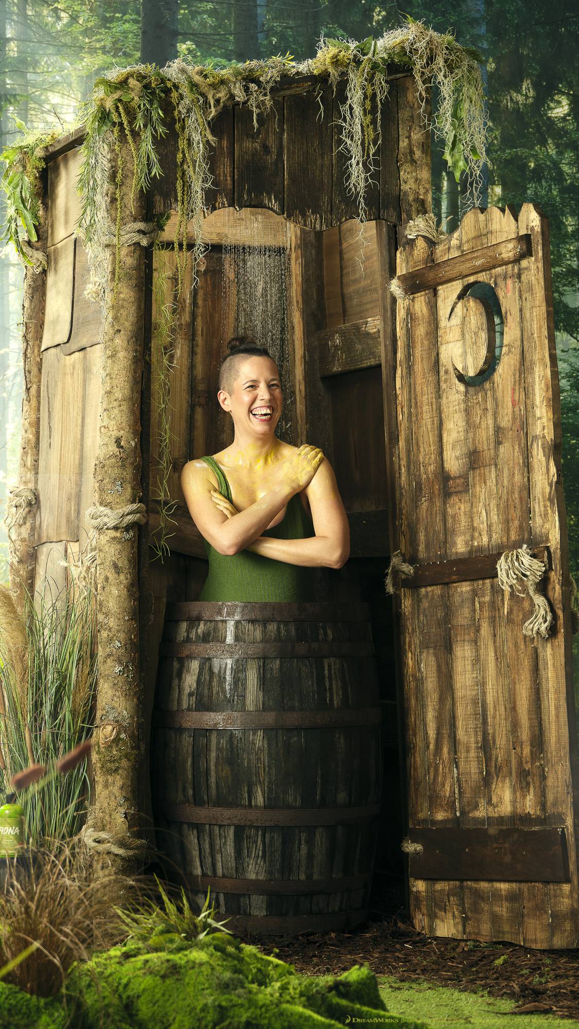 The model is in a green swimsuit while showering with the shower gel in a wooden barrel inside the iconic wonky, wooden outhouse from the Shrek films. It is surrounded by forest trees and moss. 