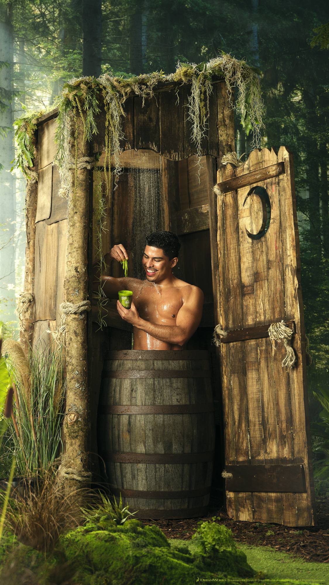 The model is holding the product pot while showering in a wooden barrel inside the iconic wonky, wooden outhouse from the Shrek films. It is surrounded by forest trees and moss. 