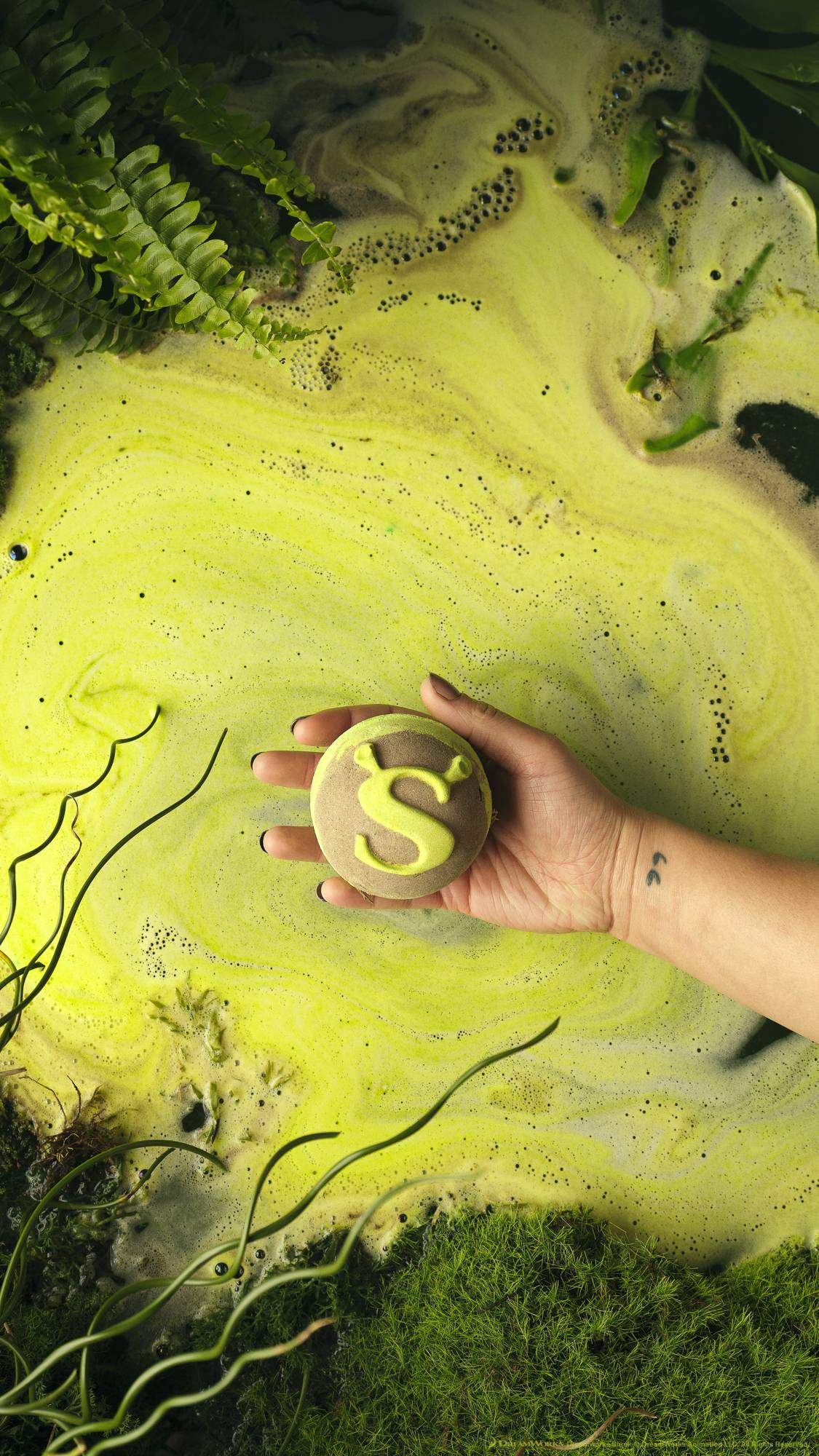 The image shows the model's forearm and hand as they hold the Shrek Swamp bath bomb just above the neon-green, swirling, foamy water below. Moss and leaf foliage surround the border. 