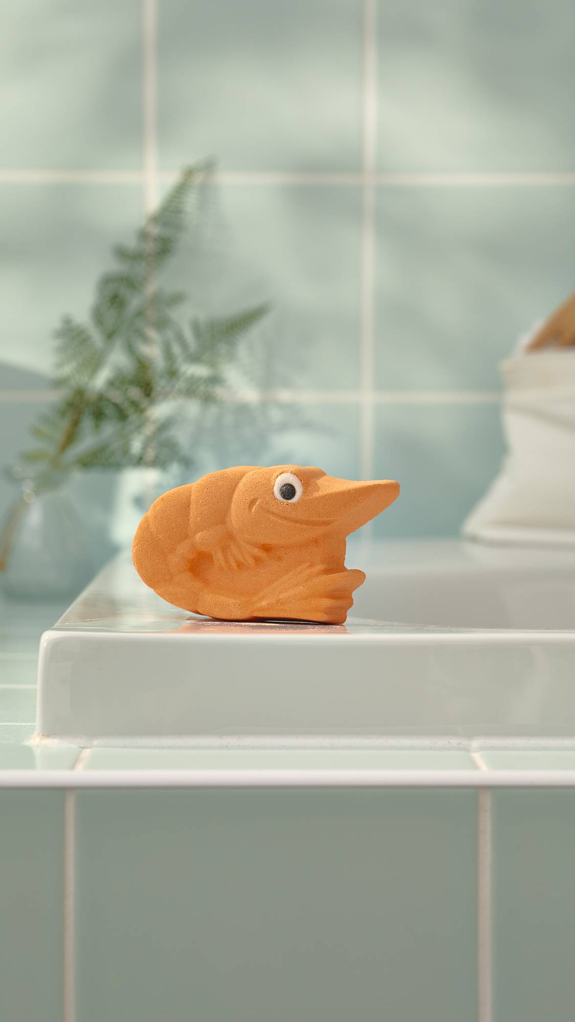The image shows the orange, Shrimp-shaped "Shrimply The Best" bath bomb sitting on the edge of a white ceramic bathtub. The background features pale mint green wall tiles and a soft green fern in a glass vase.