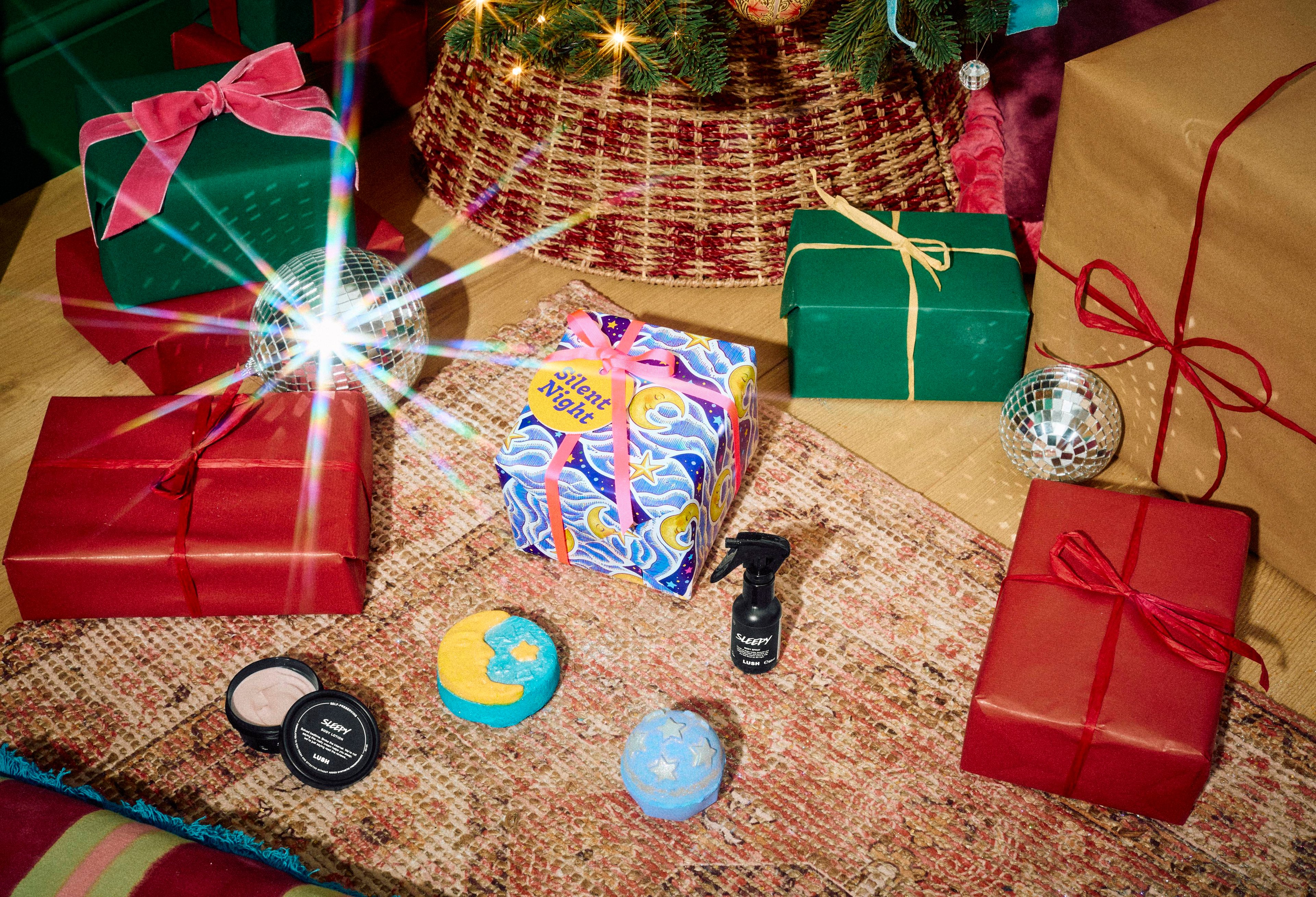 The Lush Silent Night gift box rests on a patterned rug with Sleepy body spray, Sleepy body lotion, a blue starry bath bomb, and a crescent moon bubble bar, surrounded by wrapped presents beneath a decorated Christmas tree.