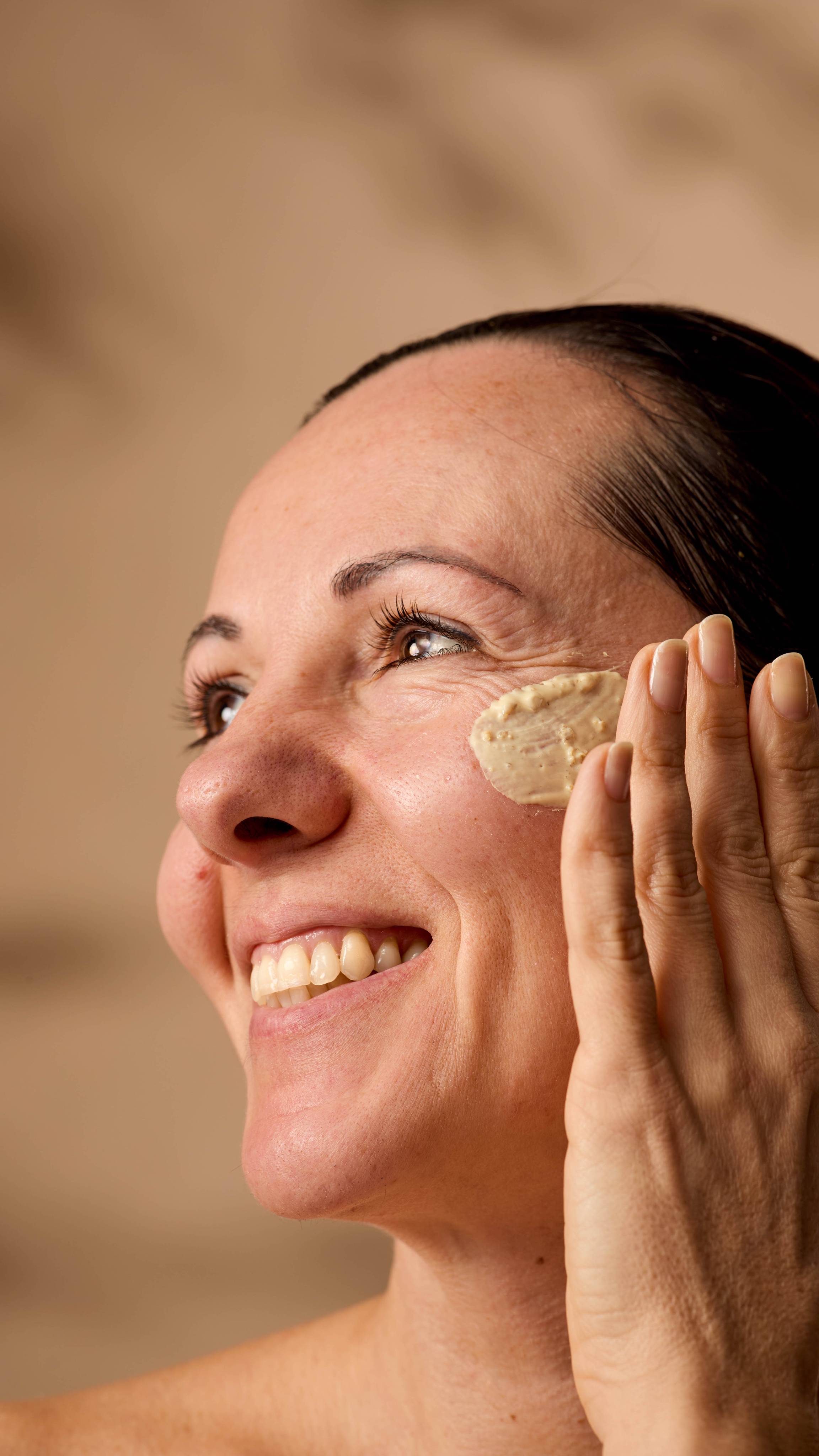 The image shows the model smiling into the distance as they apply the Skin Soothing Porridge face mask to their cheek. 