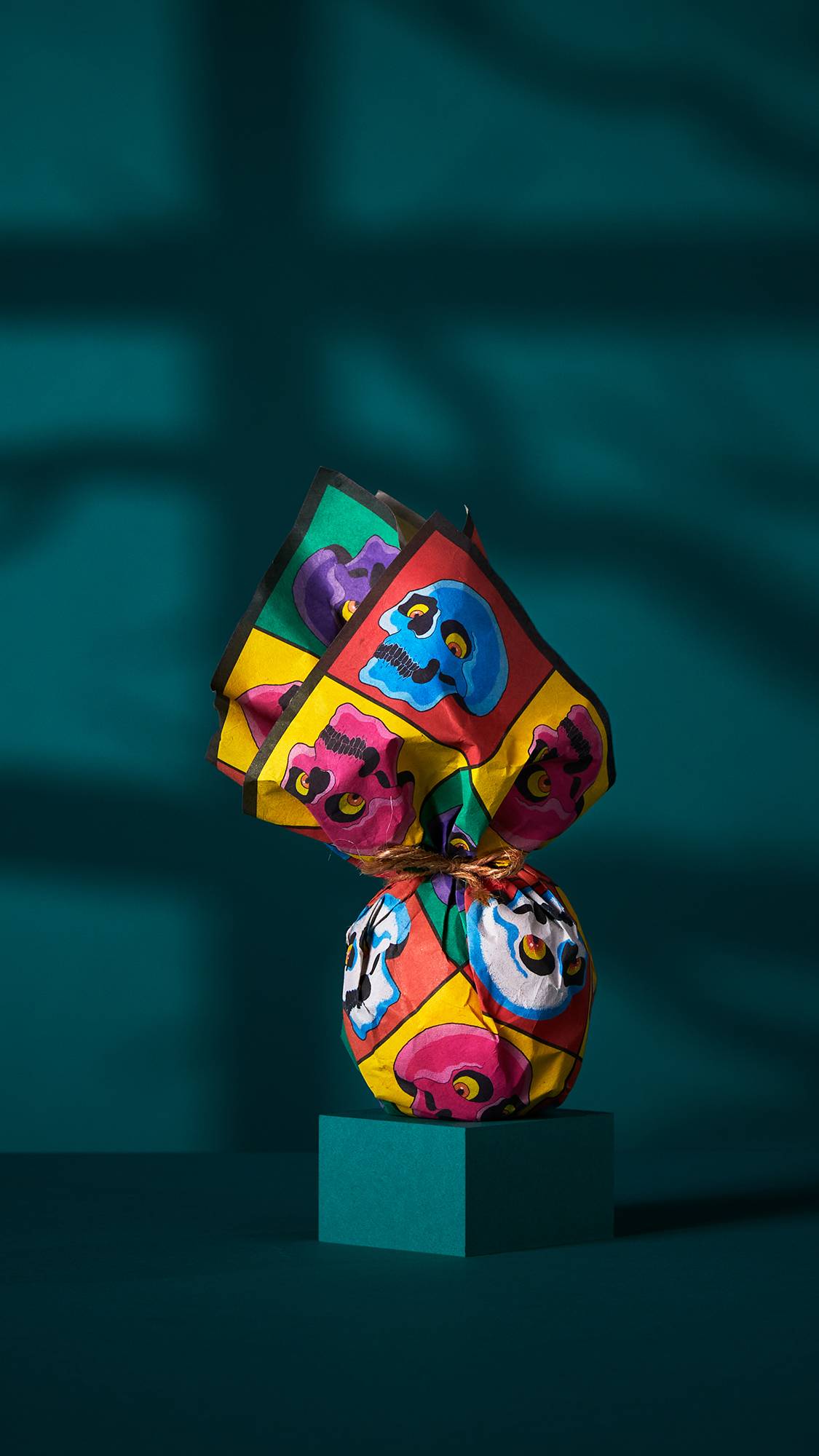 The image shows a spherical gift wrapped in a 'Skullywood' lokta paper. The paper, with its multi-coloured grid of cartoonish skulls, is tied at the top and rests on a dark teal plinth against a shadowed background.