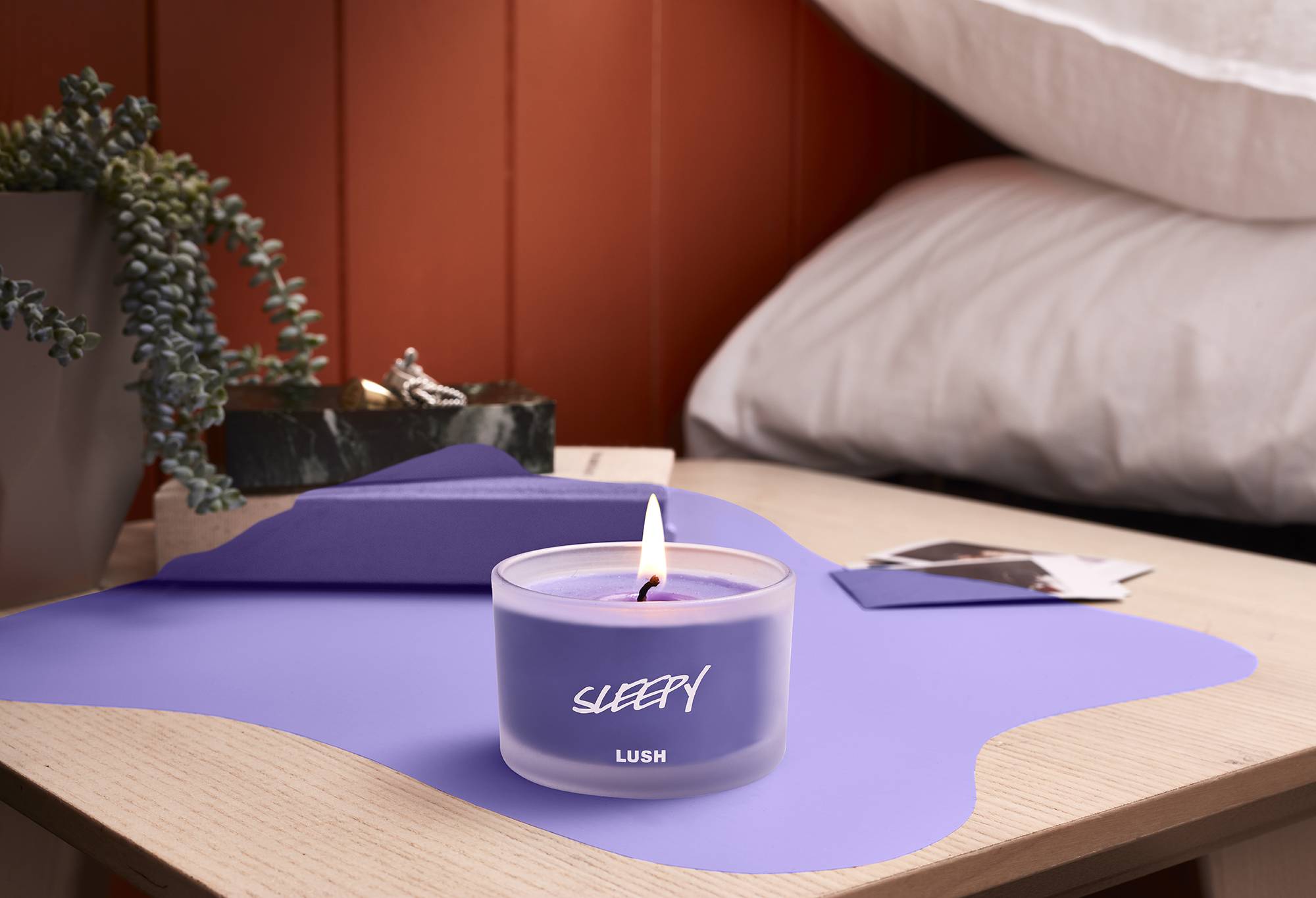 The candle is shown on a wooden bedside table, that's partly coloured the same shade of purple as the candle's wax.