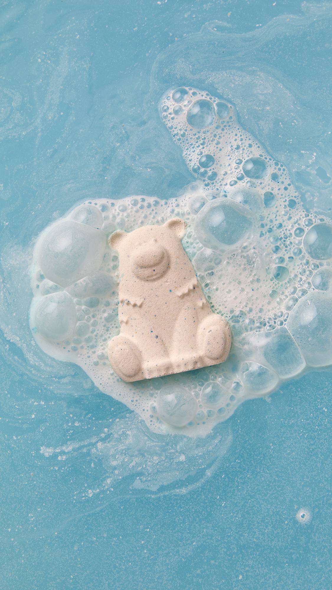 An aerial shot of the Lush Sleepy Bear bath bomb sitting on the surface of clear, blue water as it begins to dissolve giving off creamy bubbles with flecks of blue. 