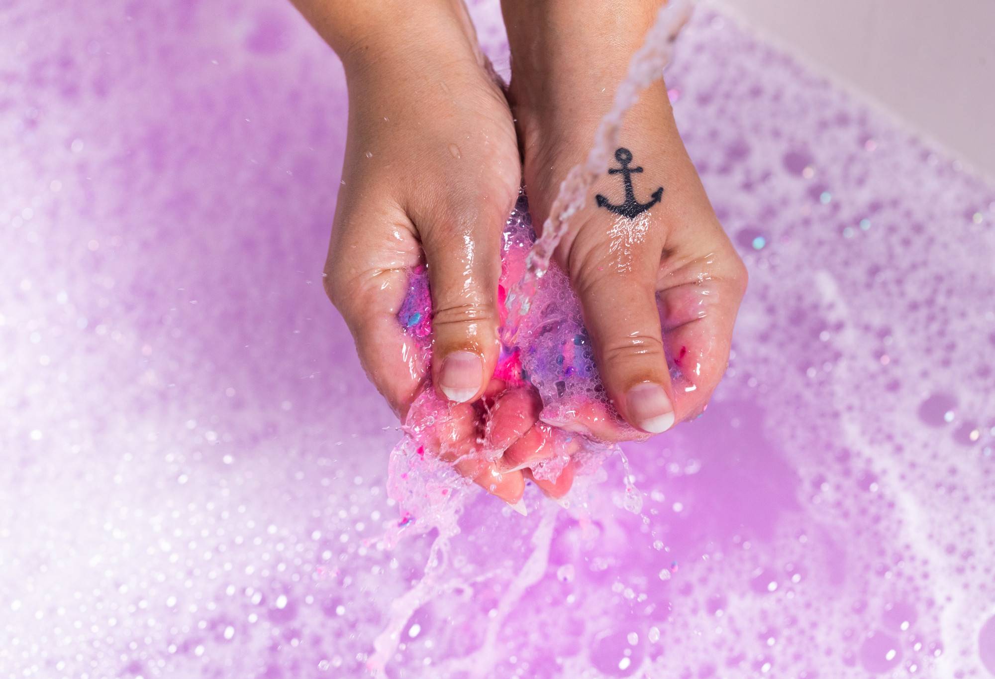 A hand with a tattoo has crumbled the pink-purple Sleepy under running water. In the background purple water and bubbles form.