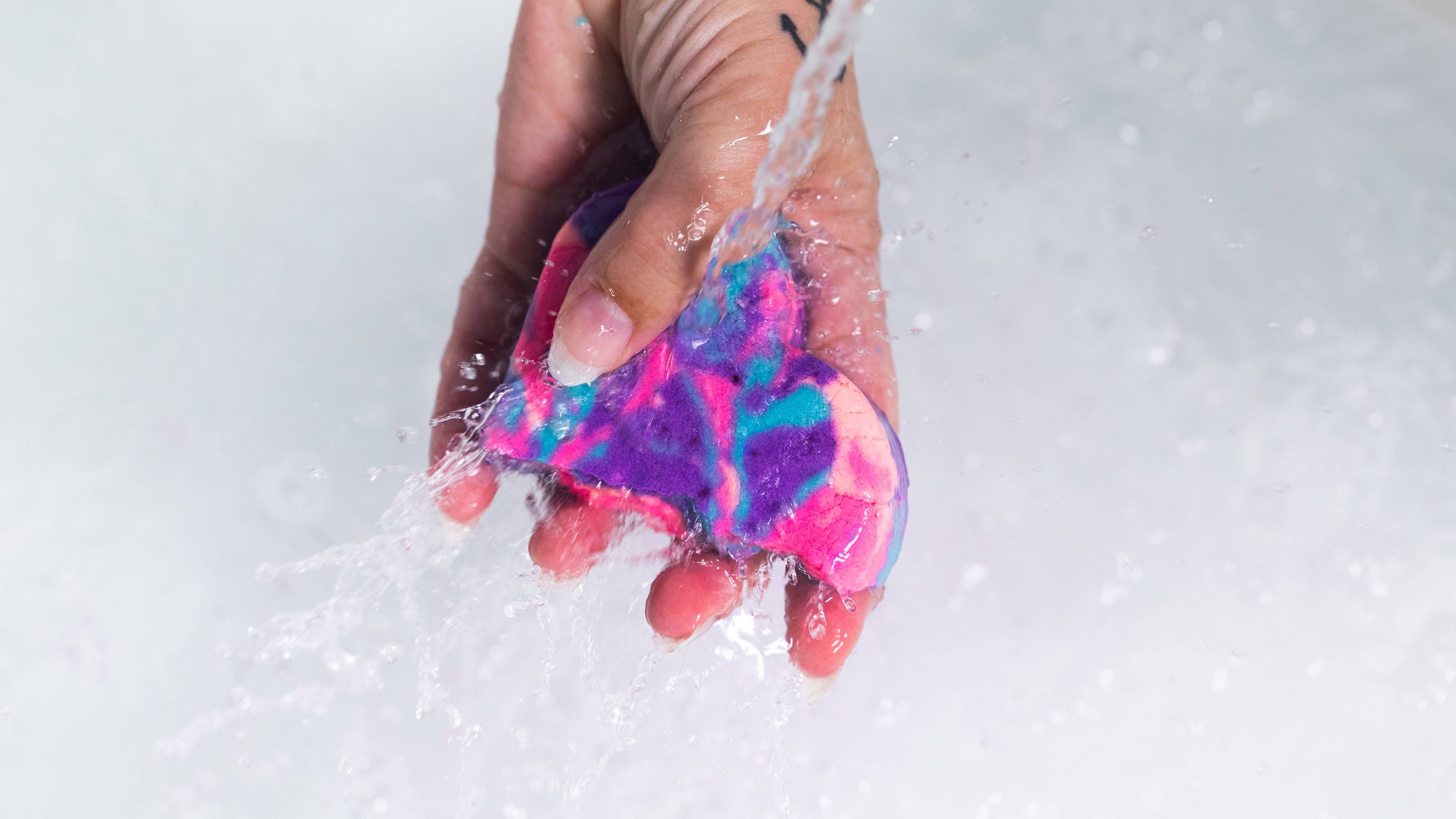 Model's hand holds half a Sleepy bubble bar, with swirling pinks, purples and blues, under running water.